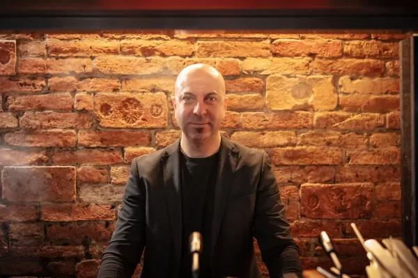 Ein Mann mit kurzen Haaren, weißem Hemd, Krawatte und dunkler Weste sitzt an einem Tisch in einem Restaurant.