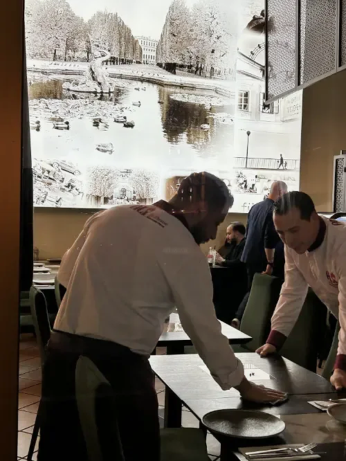 Zwei Köche in einem Restaurant bereiten einen Tisch vor, im Hintergrund ein großes Fenster mit Blick auf eine Stadt mit Wasser und Bäumen.