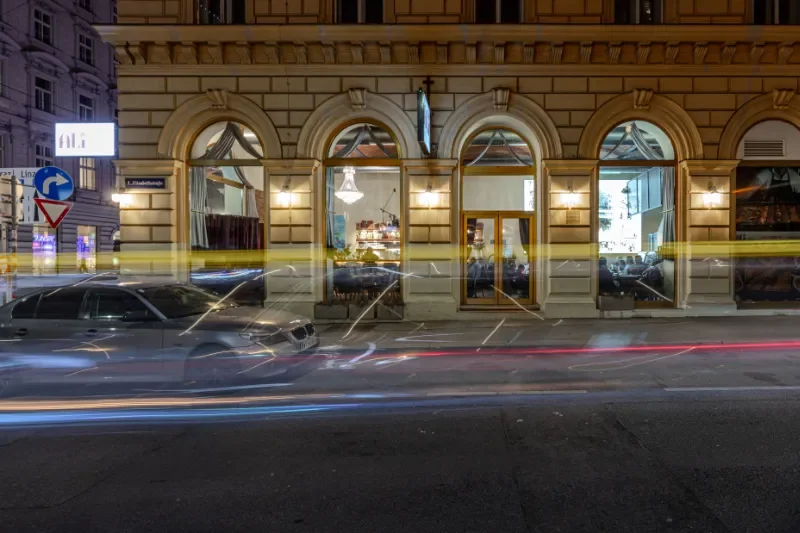 Nachtaufnahme des eleganten Restaurants Ali Grillhouse in der Operngasse 14 in 1010 Wien. Mit großen Fenstern und Innenbeleuchtung, davor stehendes Auto, Straßenbeleuchtung und Lichtspuren von vorbeifahrenden Fahrzeugen.