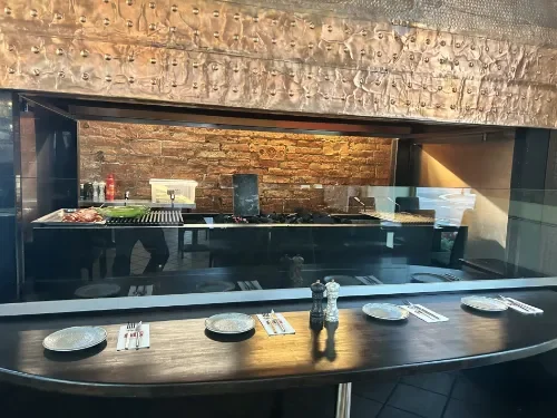 Restaurant Tisch vor dem Holzkohlengrill im Restaurant Ali Grillhouse mit einem dunklen Holztisch, sechs Serviertellern, Salz- und Pfeffermühlen, Besteck.