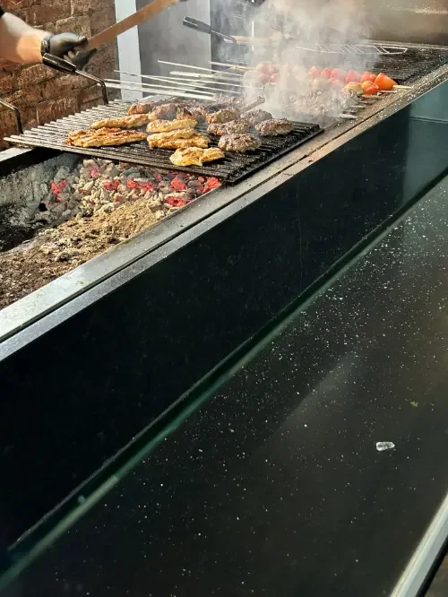 Grill mit Fleisch, Gemüse und Spieße, im Hintergrund Rauch und glühende Kohlen.