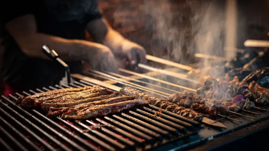 Ali Grillhouse Holzkohlengrill mit verschiedenen Fleischsorten die gerade gegrillt werden, Rauch steigt auf undHände mit Grill-Zange sind zu sehen