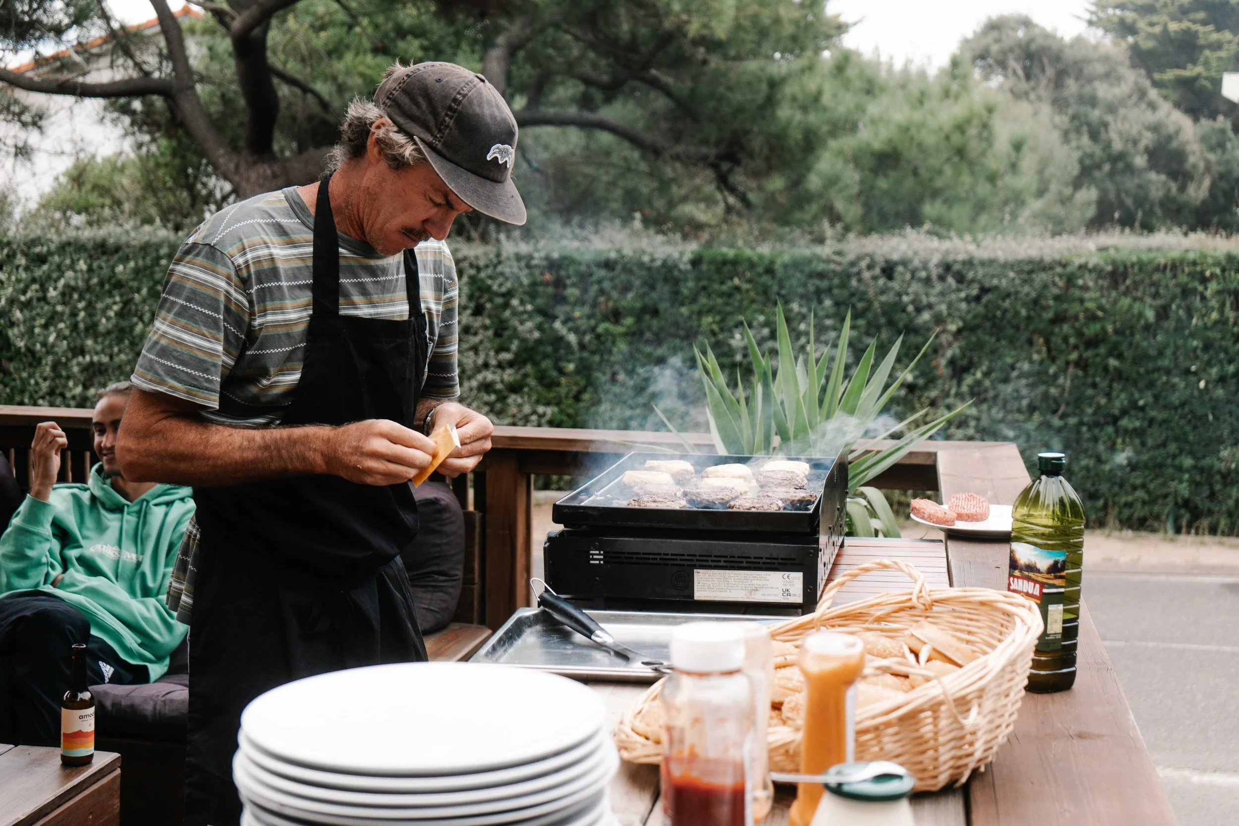 Man cooking hamburgers on a grill outdoors at a gathering, with a woman sitting nearby and various condiments and dishes on a wooden table.