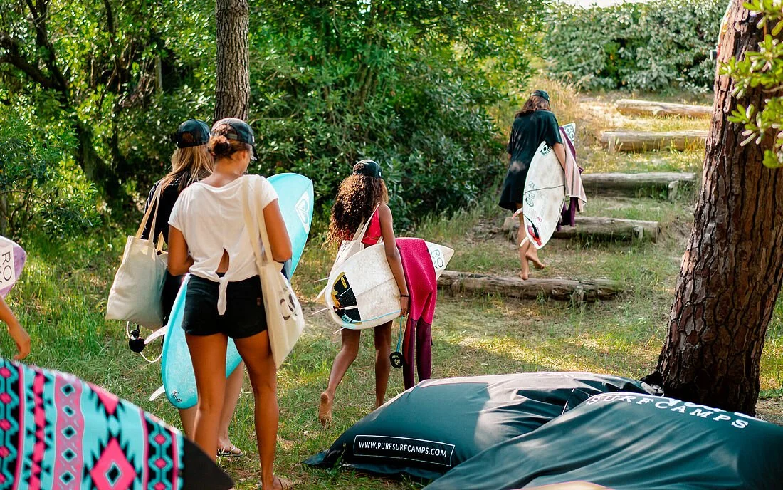 Group of young women and girls walking up wooden steps through a forested area with surfboards, wearing casual summer clothes and hats, possibly heading to surf at a nearby beach.
