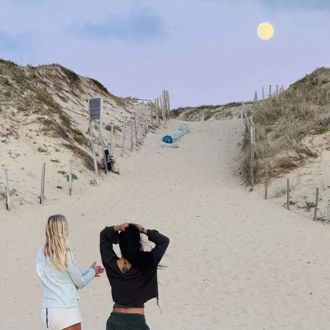 Two women standing on a sandy beach near sand dunes, looking up at the sky with a full moon, with a path leading up the dunes in the background.