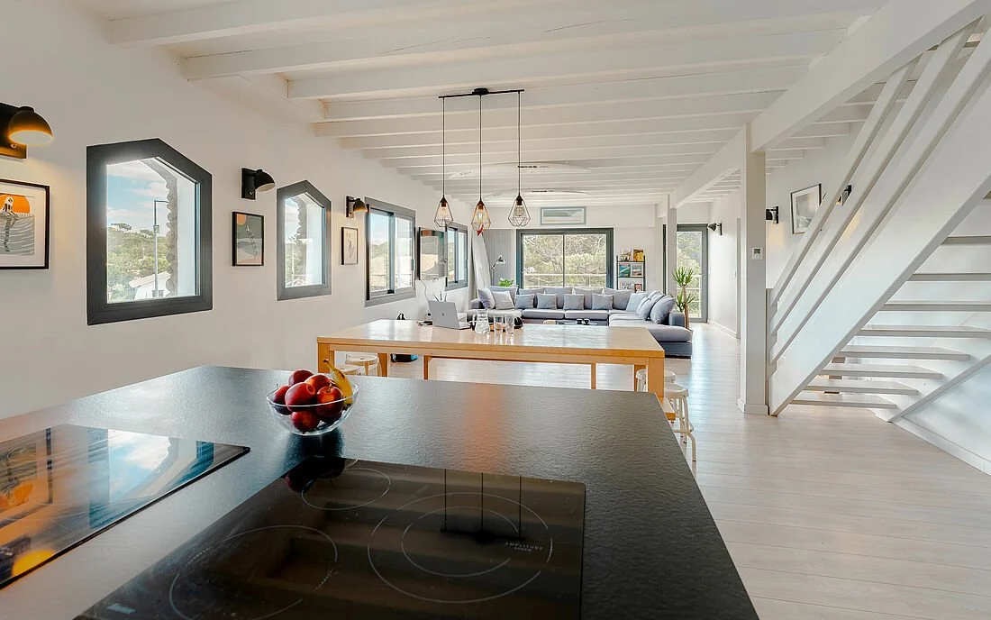 A bright open-concept living space with a kitchen island, a wooden dining table, and a spacious gray sectional sofa. Large windows let in natural light, and there are modern pendant lights hanging above the dining table.