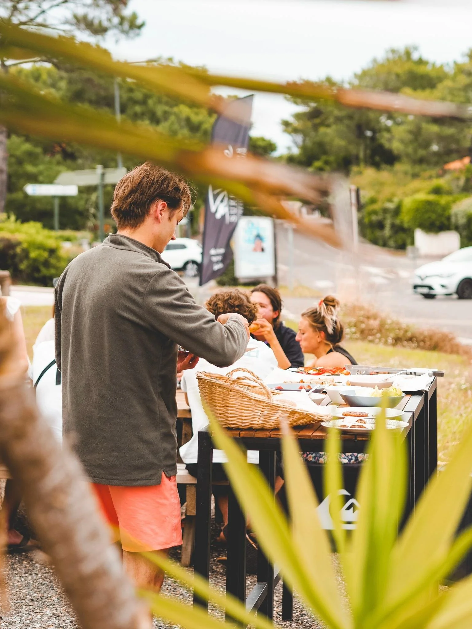 People gathering outdoors at a buffet-style table with food, with a man serving himself, surrounded by trees, cars, and flags in the background.
