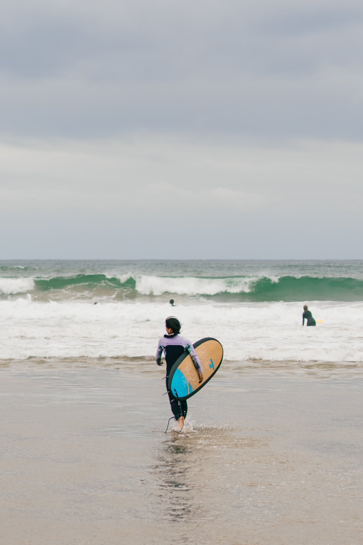 Child with a wetsuit and surfboard walking into the ocean with other surfers and waves in the background on a cloudy day.