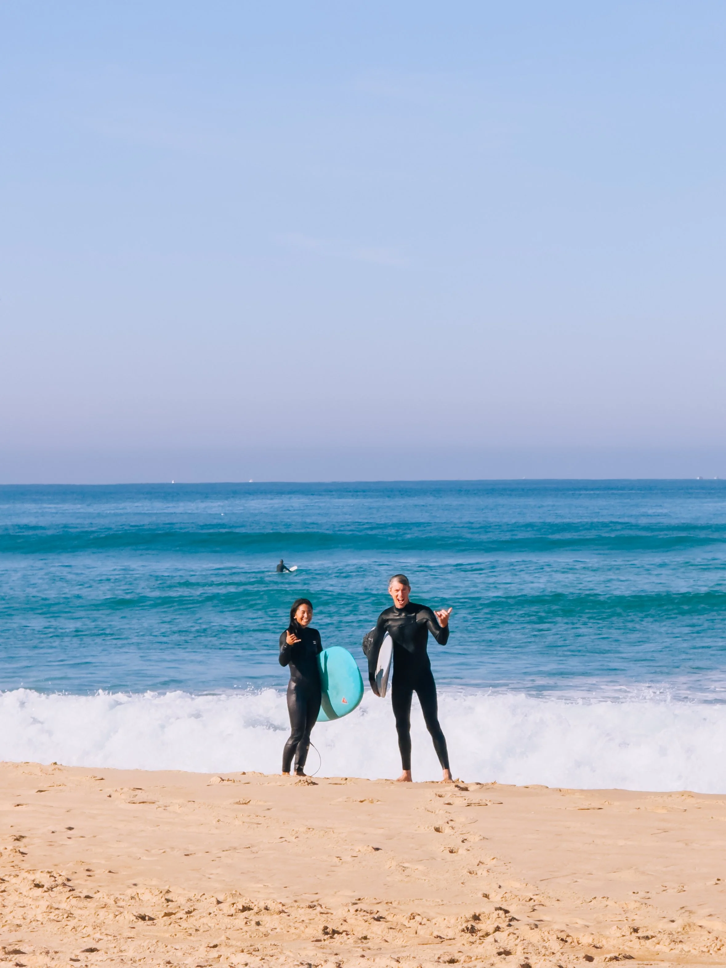 Two women in wetsuits standing on the sand at the beach, holding surfboards, with the ocean and a surfer in the background.
