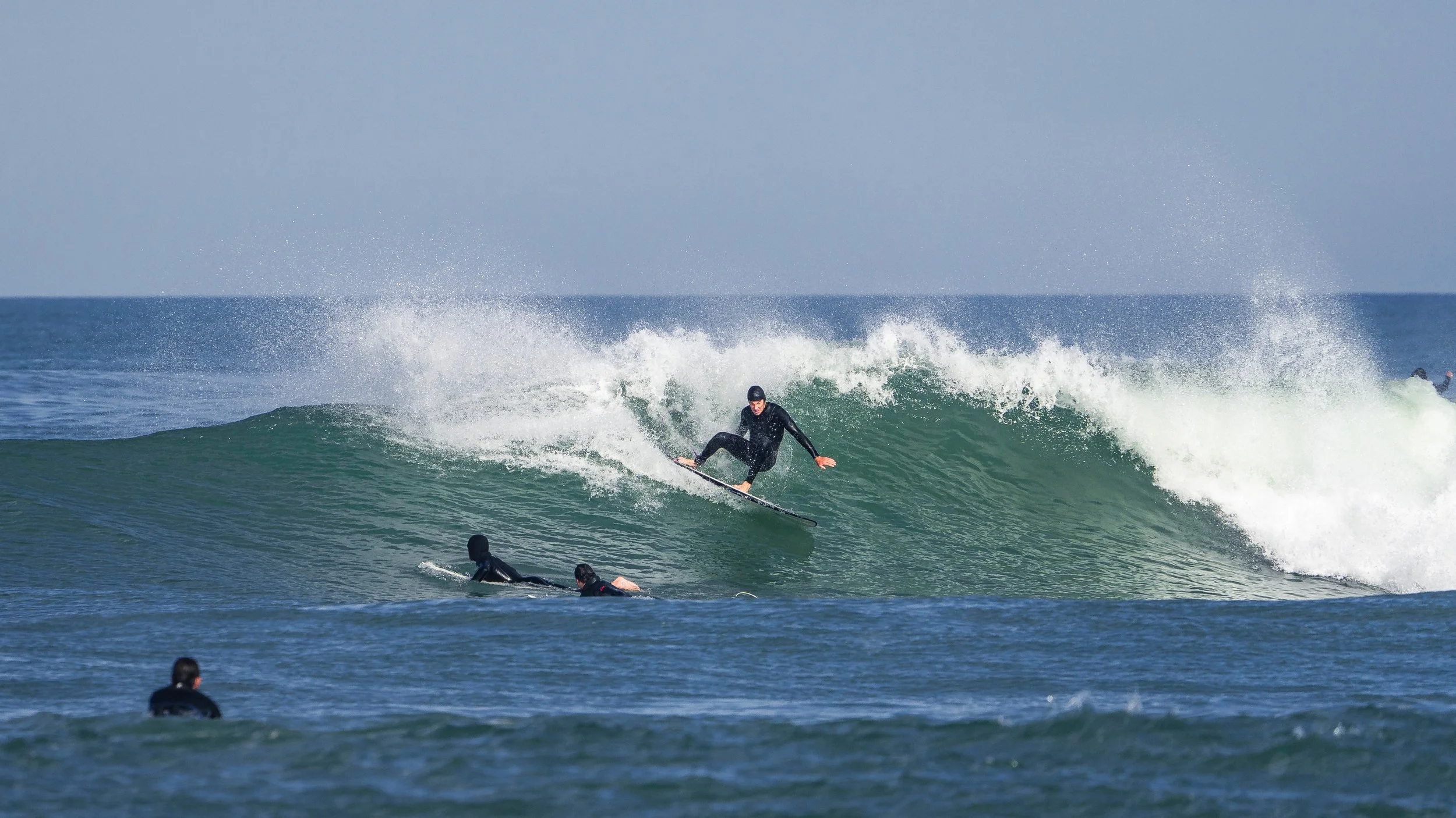 A surfer riding a wave with several other surfers in the water, under a clear sky.