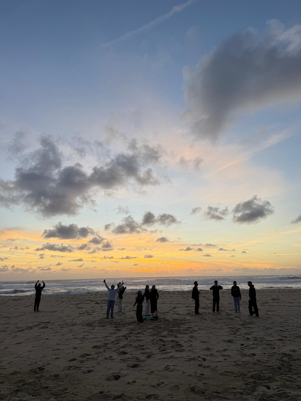 A group of people standing and sitting on a sandy beach during sunset, with some holding hands, making gestures, and facing the ocean, under a partly cloudy sky.