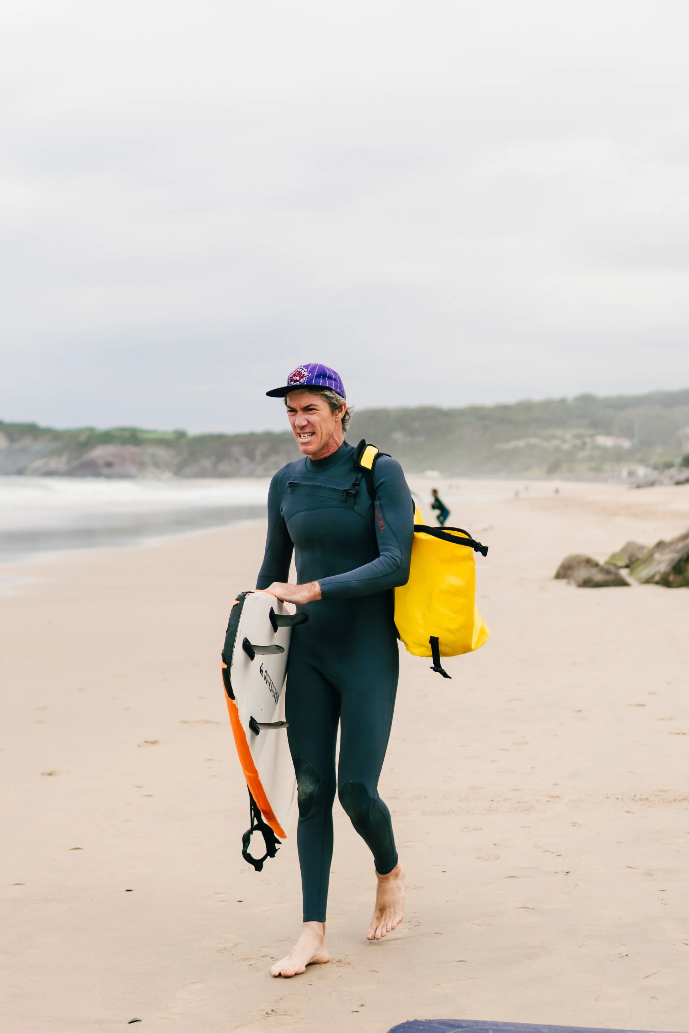 A man in a wetsuit carrying a bodyboard and a yellow dry bag walking along a sandy beach with cliffs in the background.