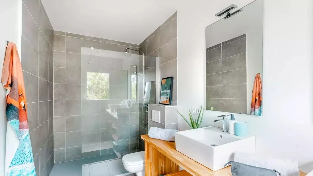 Modern bathroom with gray tiles, a walk-in shower with a glass partition, a white vessel sink on a wooden vanity, a large mirror, and a towel hanging on a wall