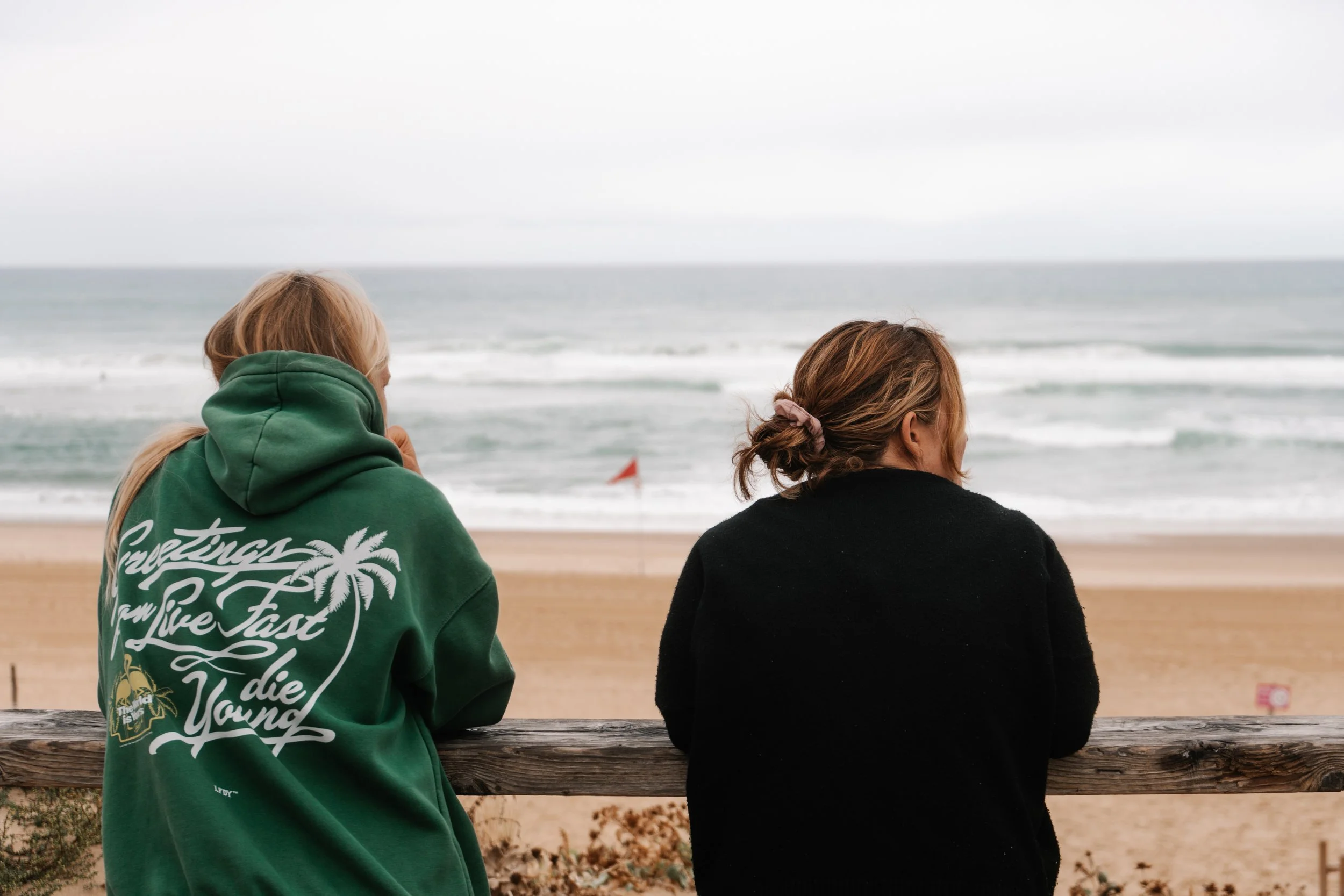 Two women sitting on a wooden railing, overlooking a beach and ocean with waves, under an overcast sky.