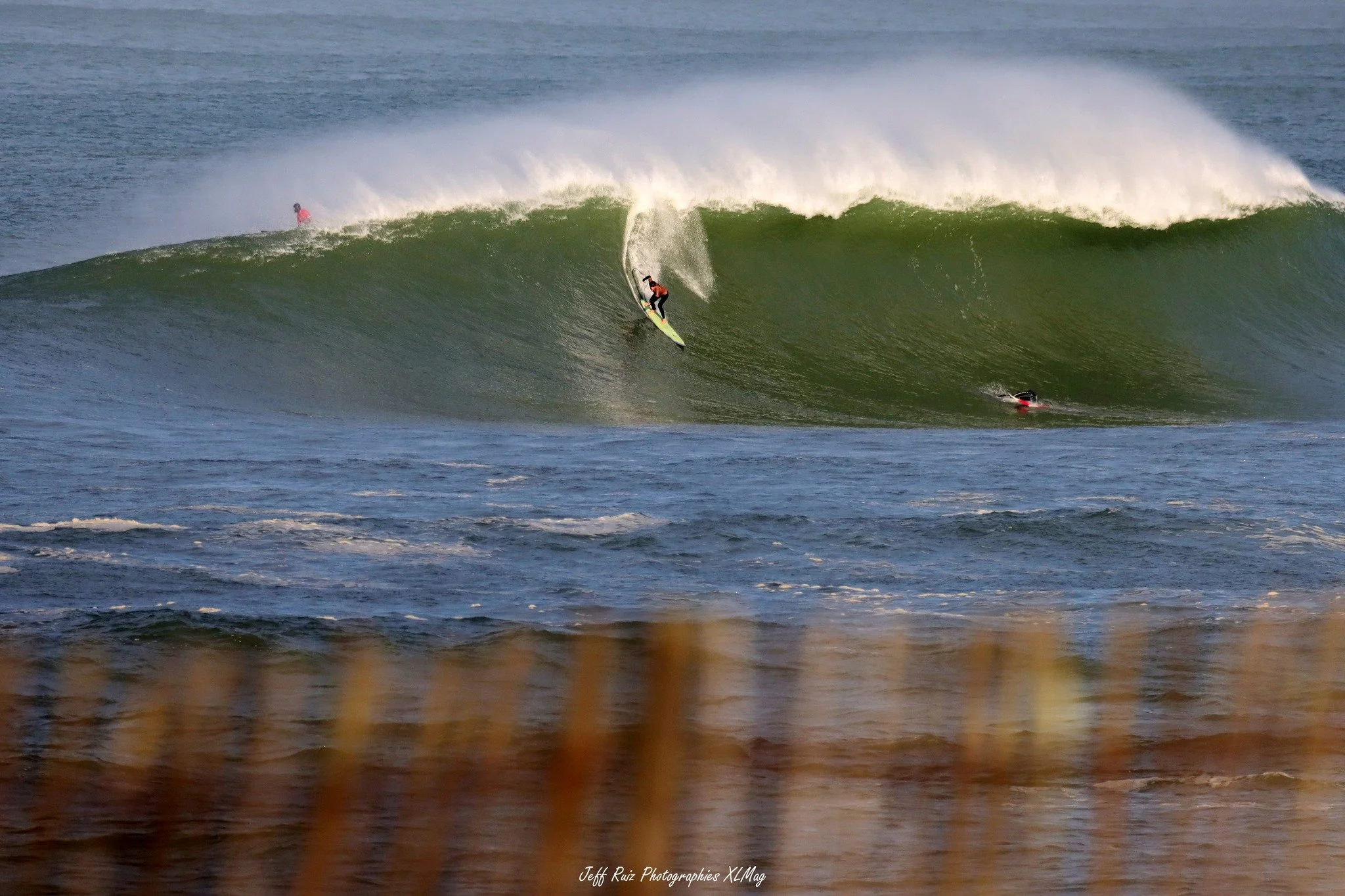 A surfer riding a large green wave with two other surfers visible in the water and a person on the shore in the background.