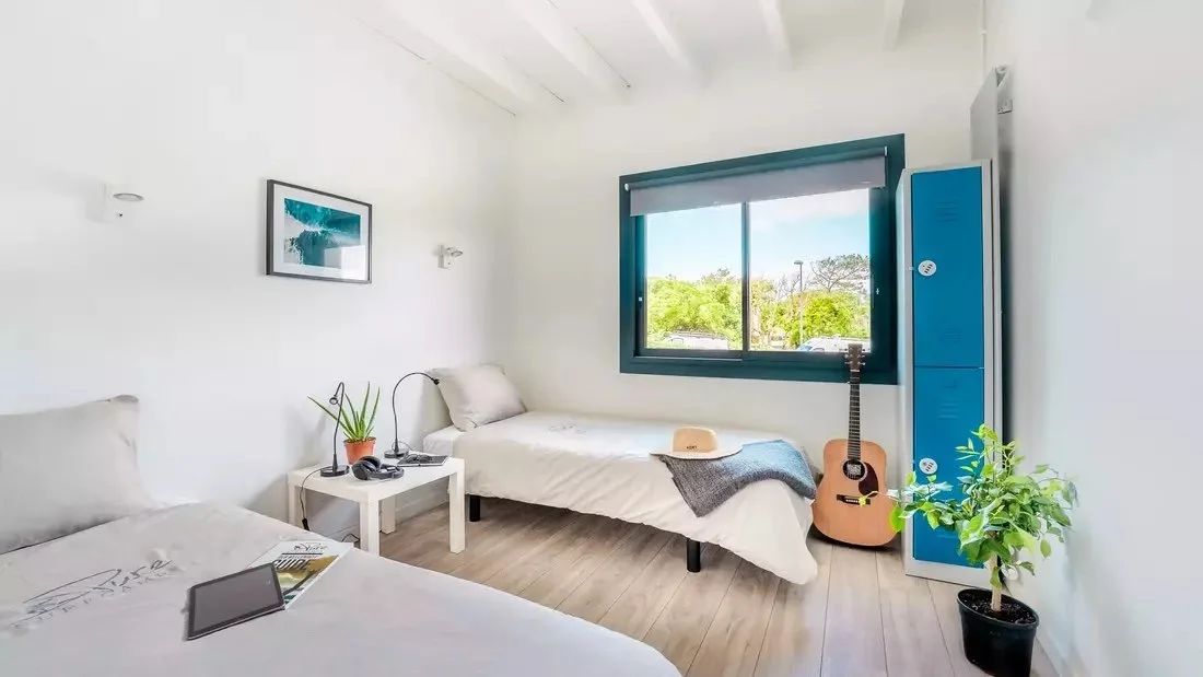 Bright bedroom with two beds, window showing trees outside, blue locker, acoustic guitar, potted plants, and a straw hat.