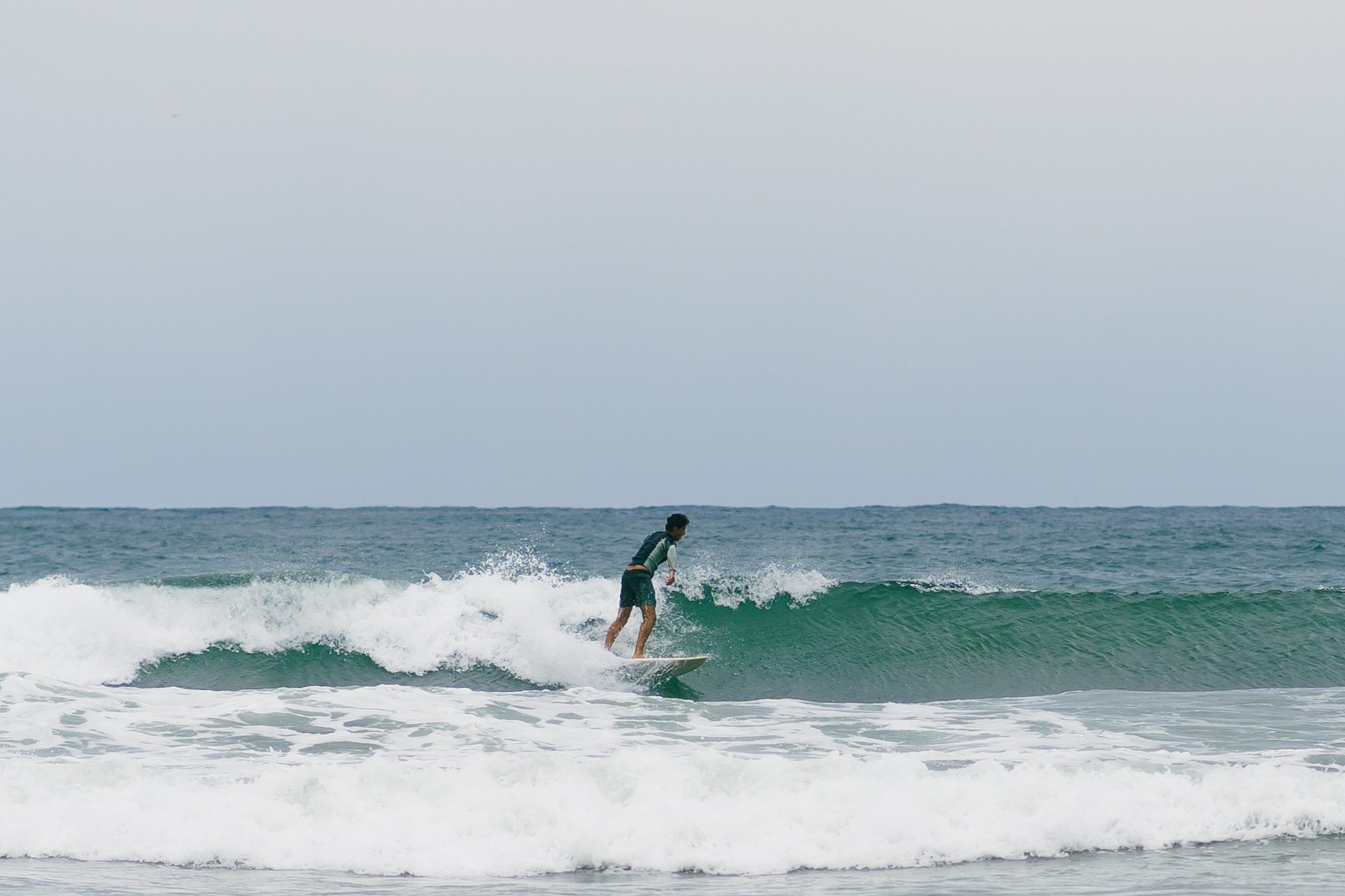 A person surfing on a small wave at the beach, wearing a green wetsuit top and black shorts.