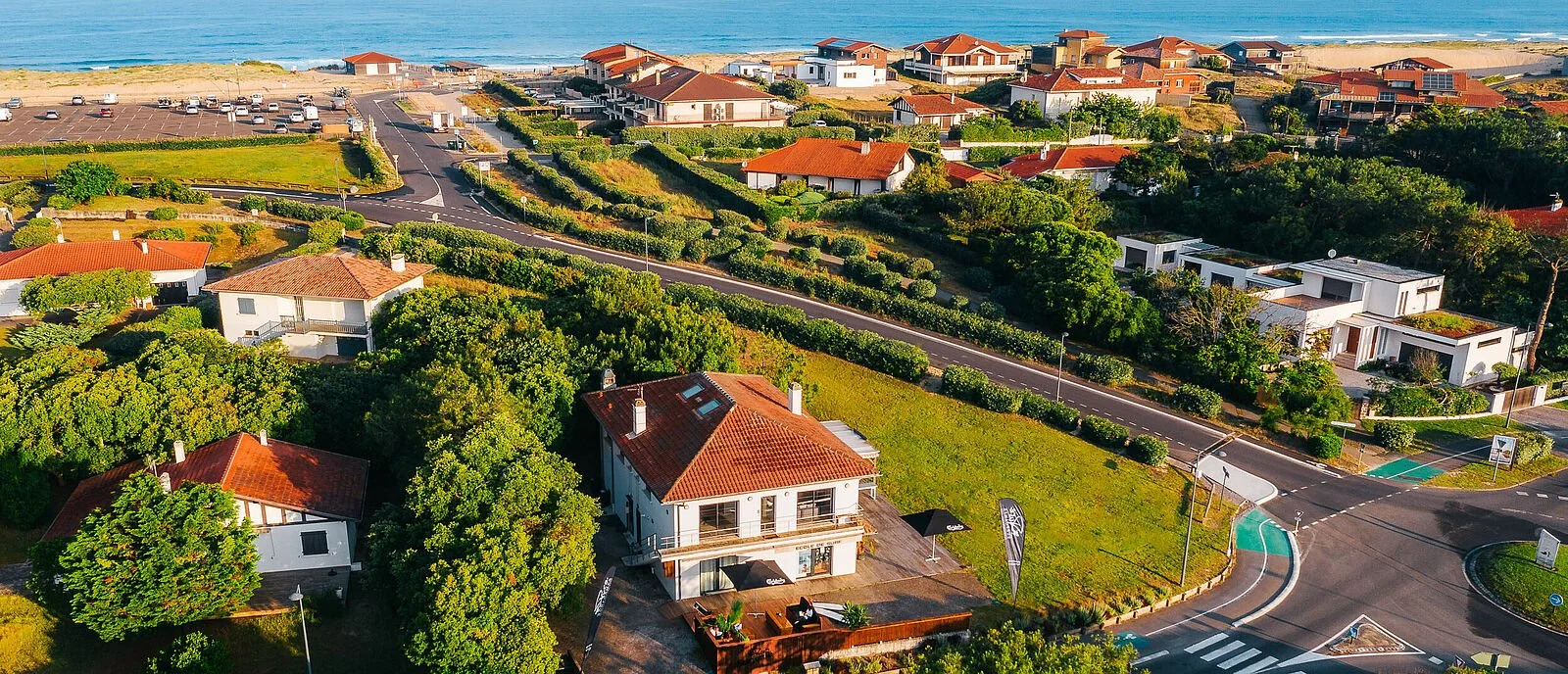 Aerial view of a coastal residential area with houses, roads, greenery, and the ocean in the background.