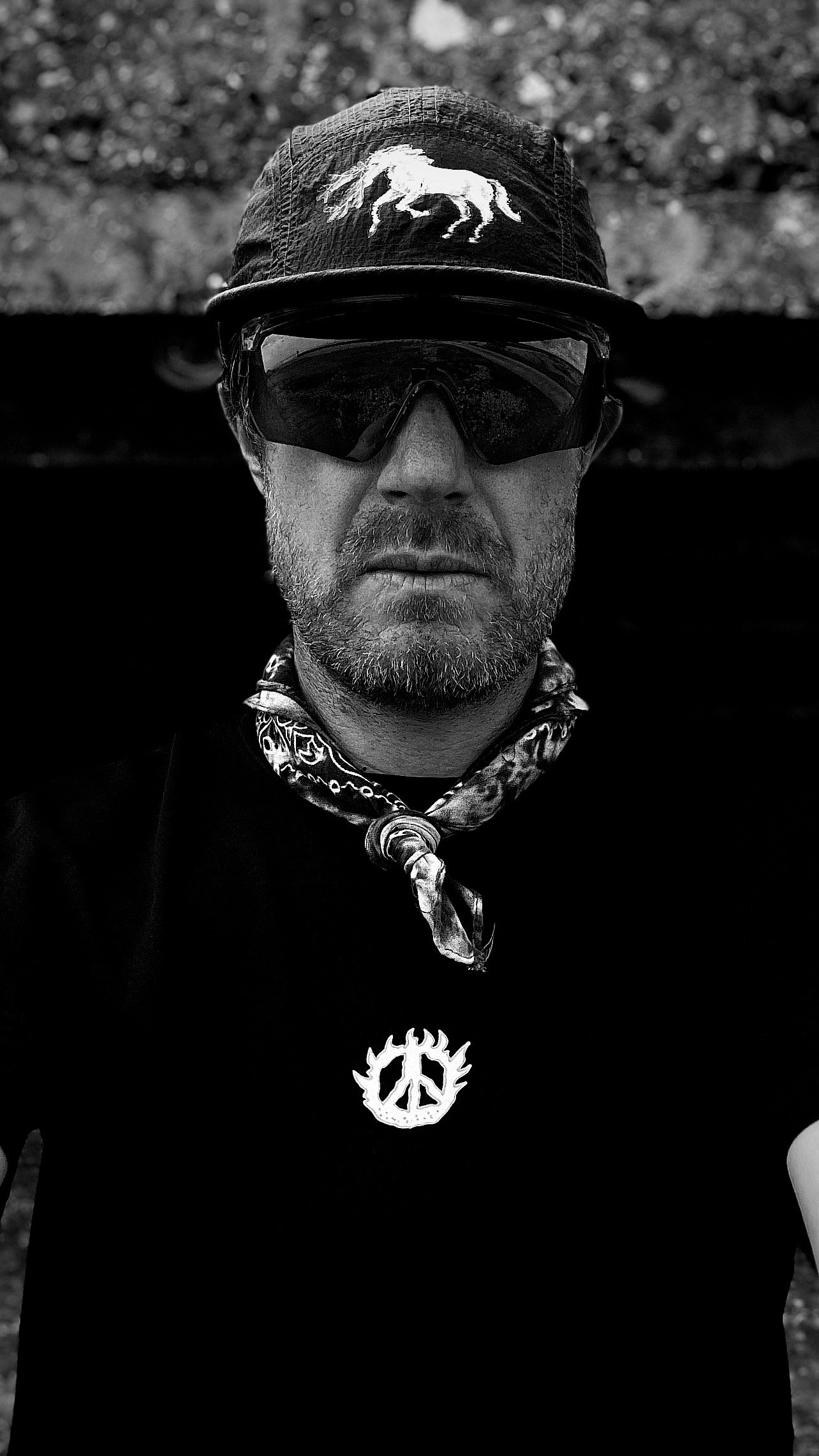 A man wearing a black cap with a horse emblem, black sunglasses, a bandana, and a black t-shirt with a peace symbol, outdoors.