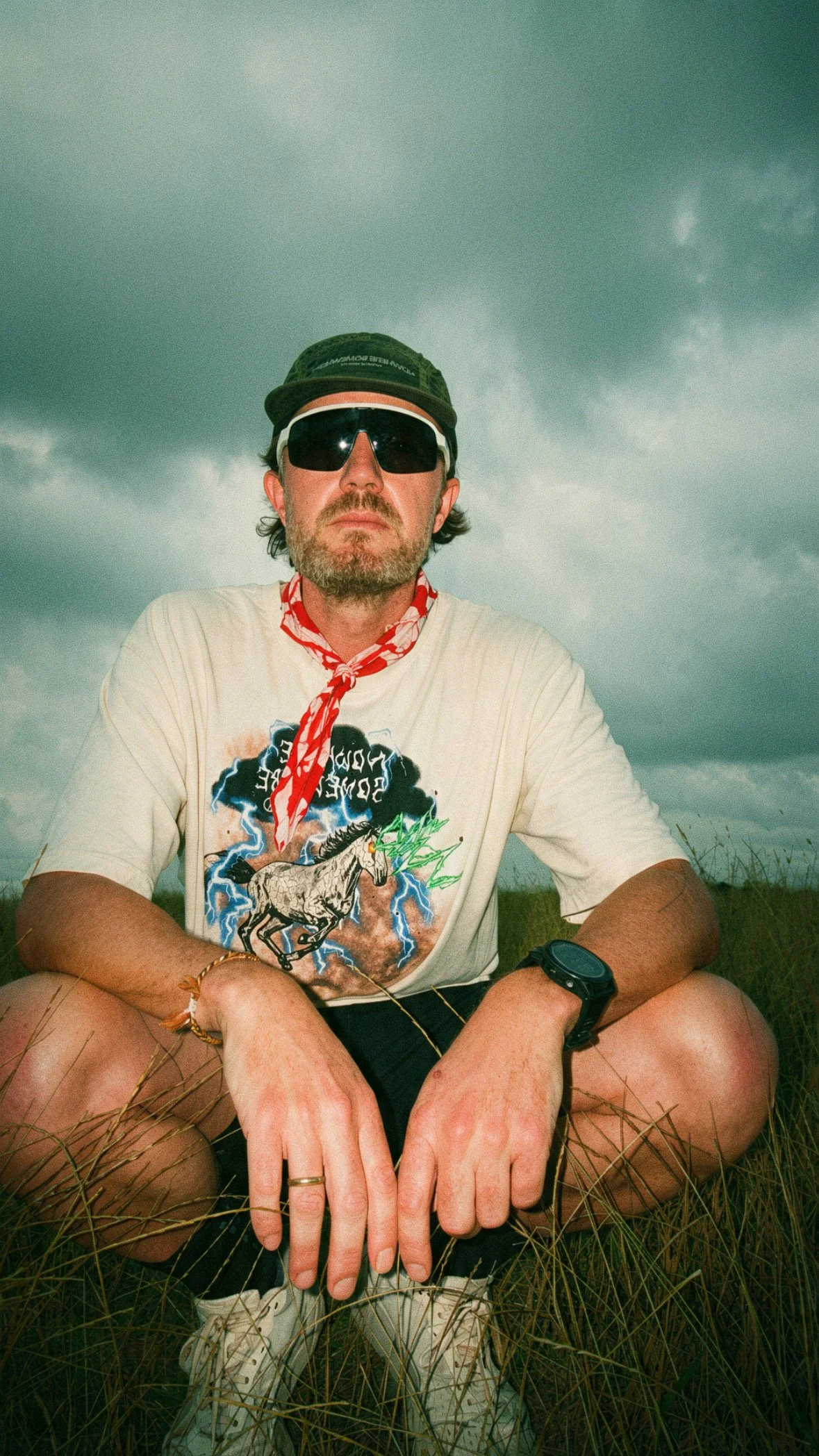 A man sitting outdoors in a field of tall grass during a cloudy day, wearing sunglasses, a green cap, a graphic T-shirt, a red bandana, a black wristwatch, and gold rings, with clouds in the sky behind him.