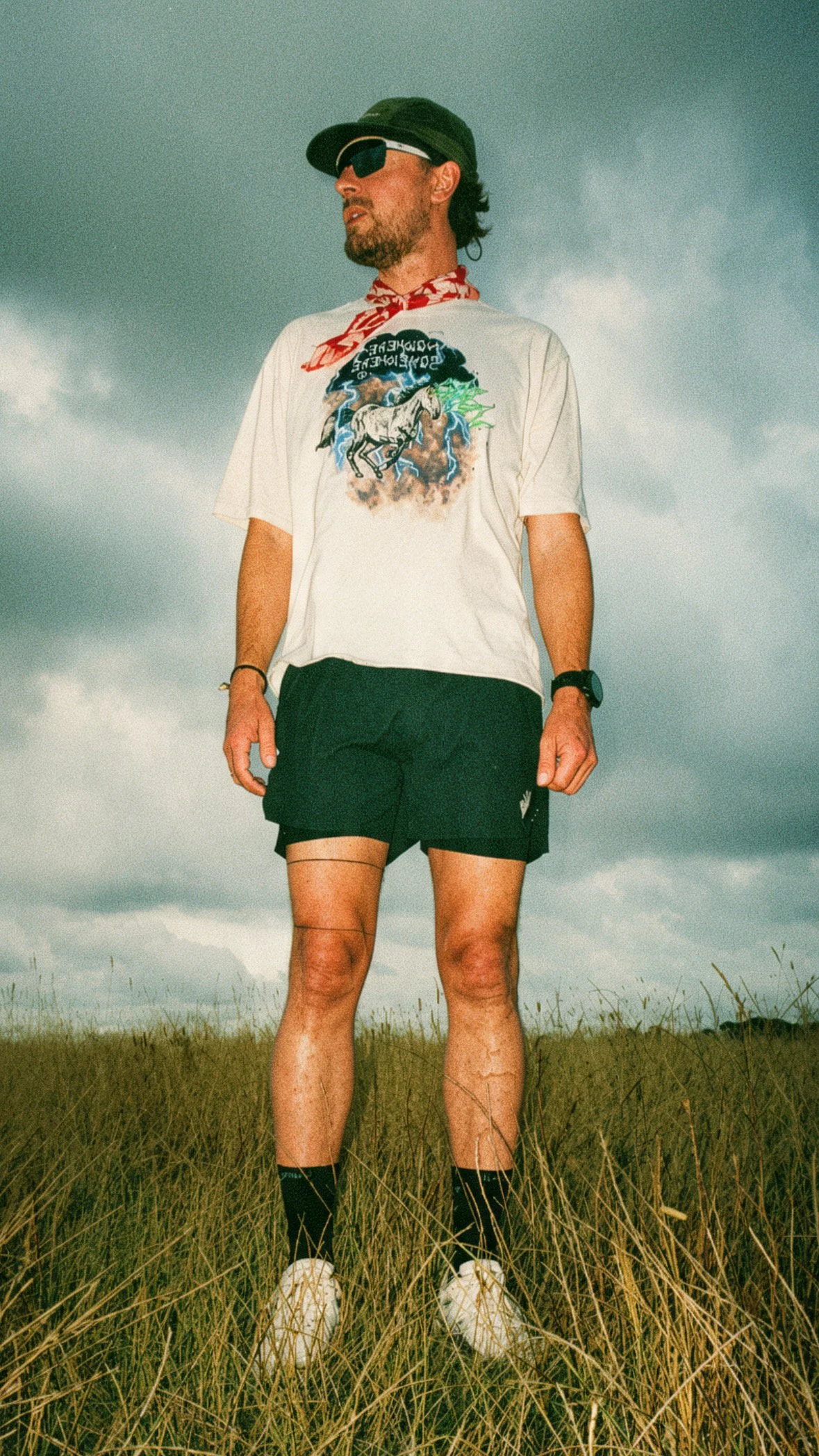 Person standing in a grassy field wearing a T-shirt with a graphic print, black shorts, sneakers, sunglasses, and a cap. The sky is cloudy.