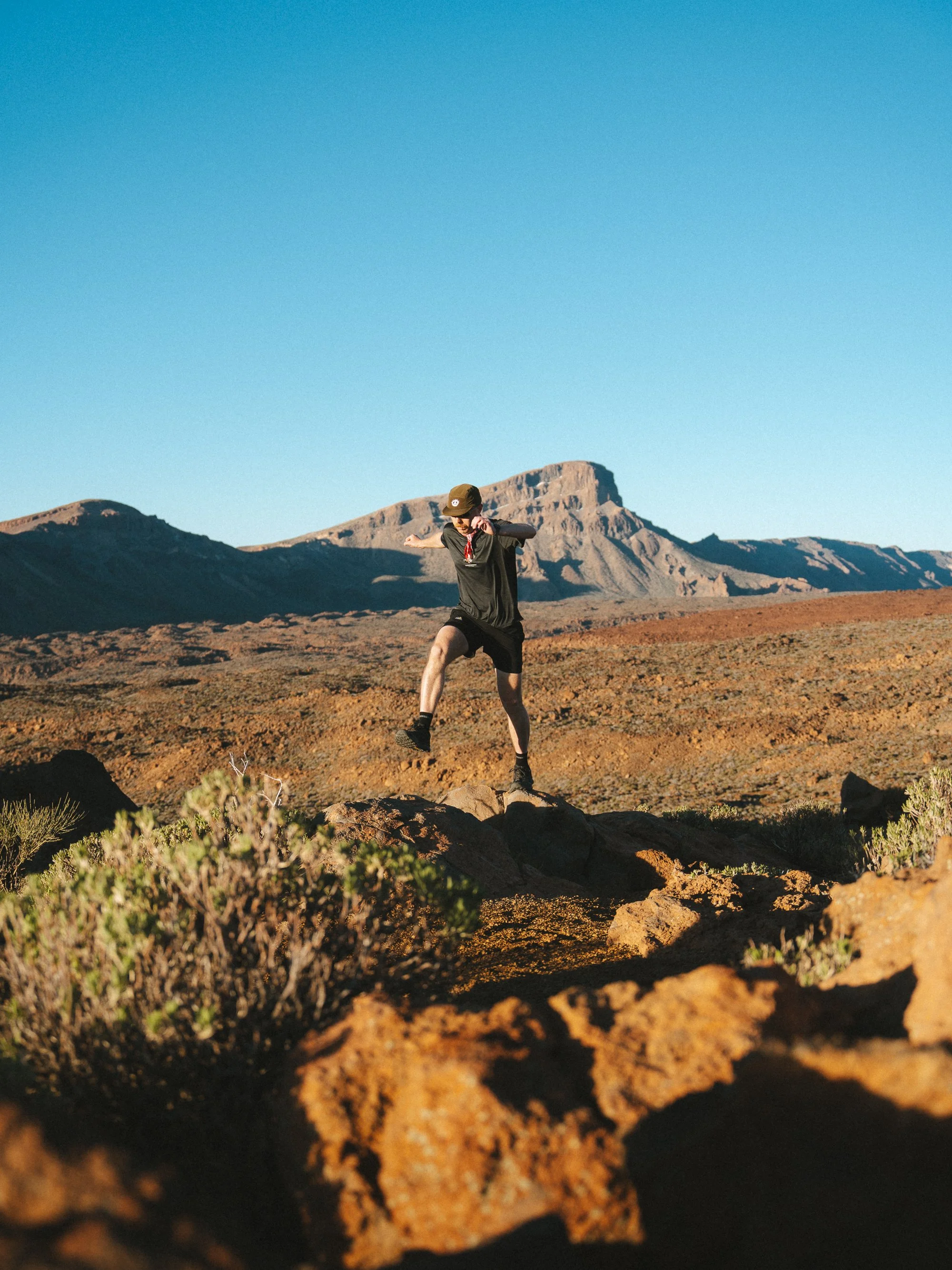 A person in black sportswear running or jumping on rocky terrain in a desert landscape with mountains in the background under a clear blue sky.