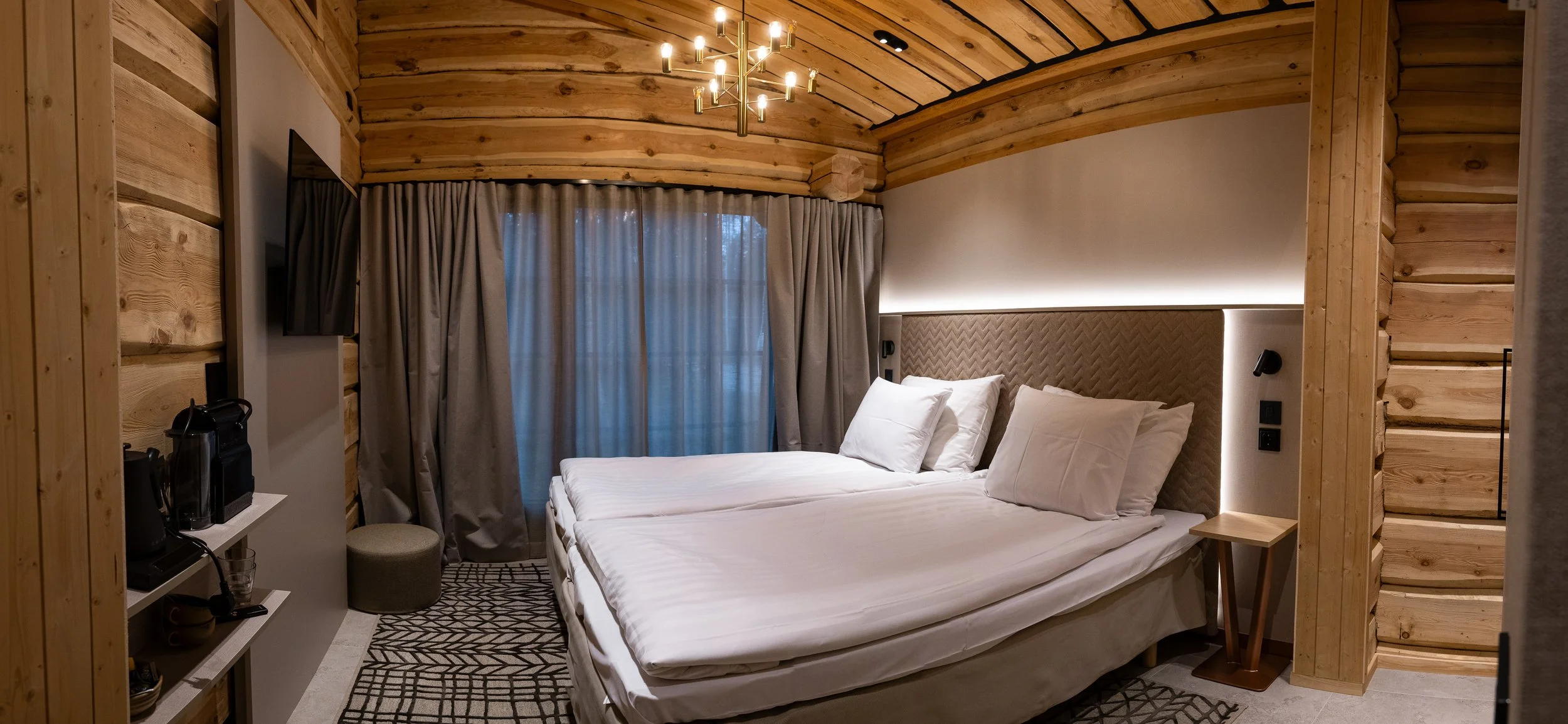 Valkoinen sänky huoneessa, jossa puiset seinät ja katto, harmaat verhot ikkunan edessä, yläpuolella kulta- tai messinkivalaisin.