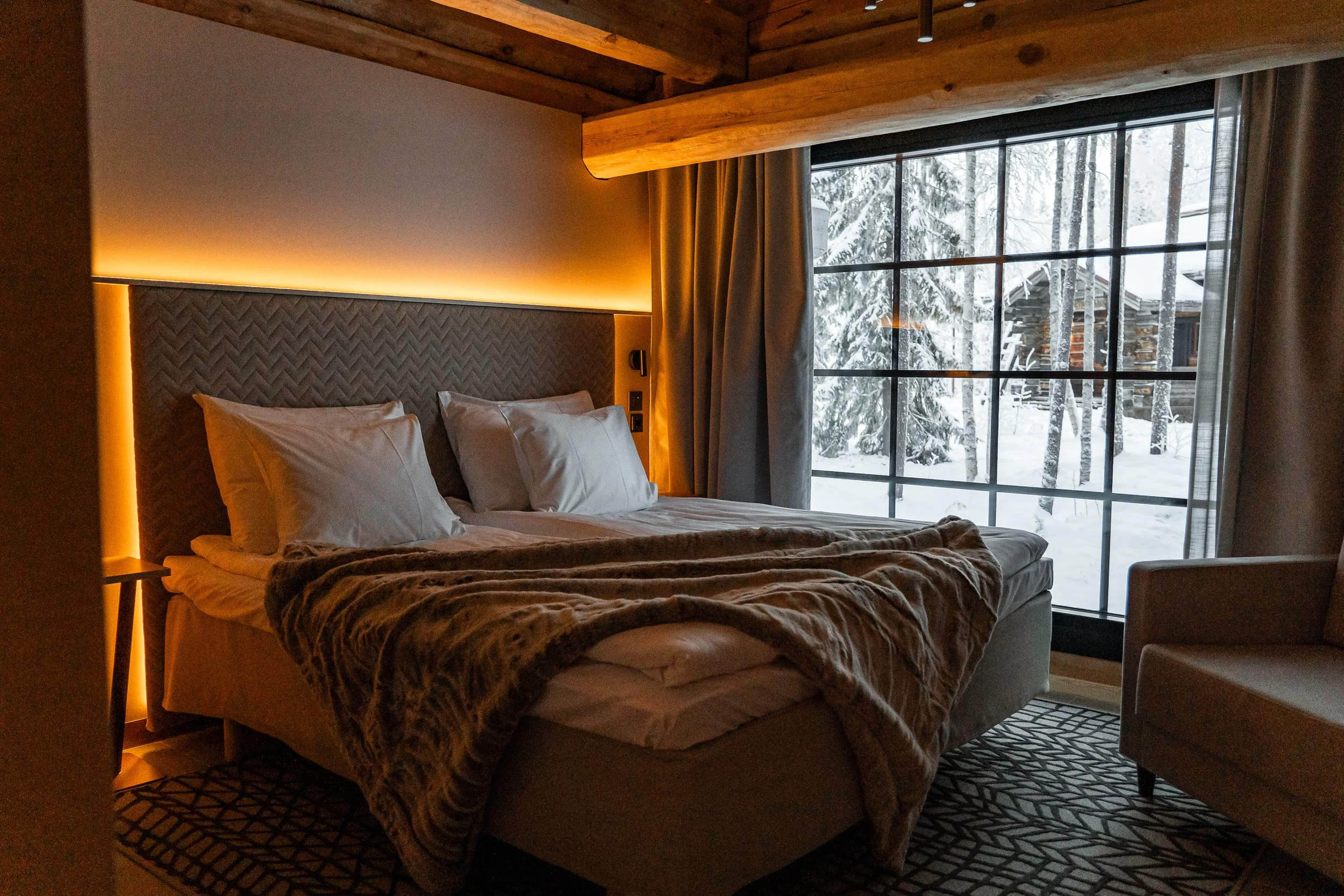 Mukava hotellisänky vuodesohvalla ja suuri ikkuna, josta näkee lumisen metsän ja mökin pihalle.