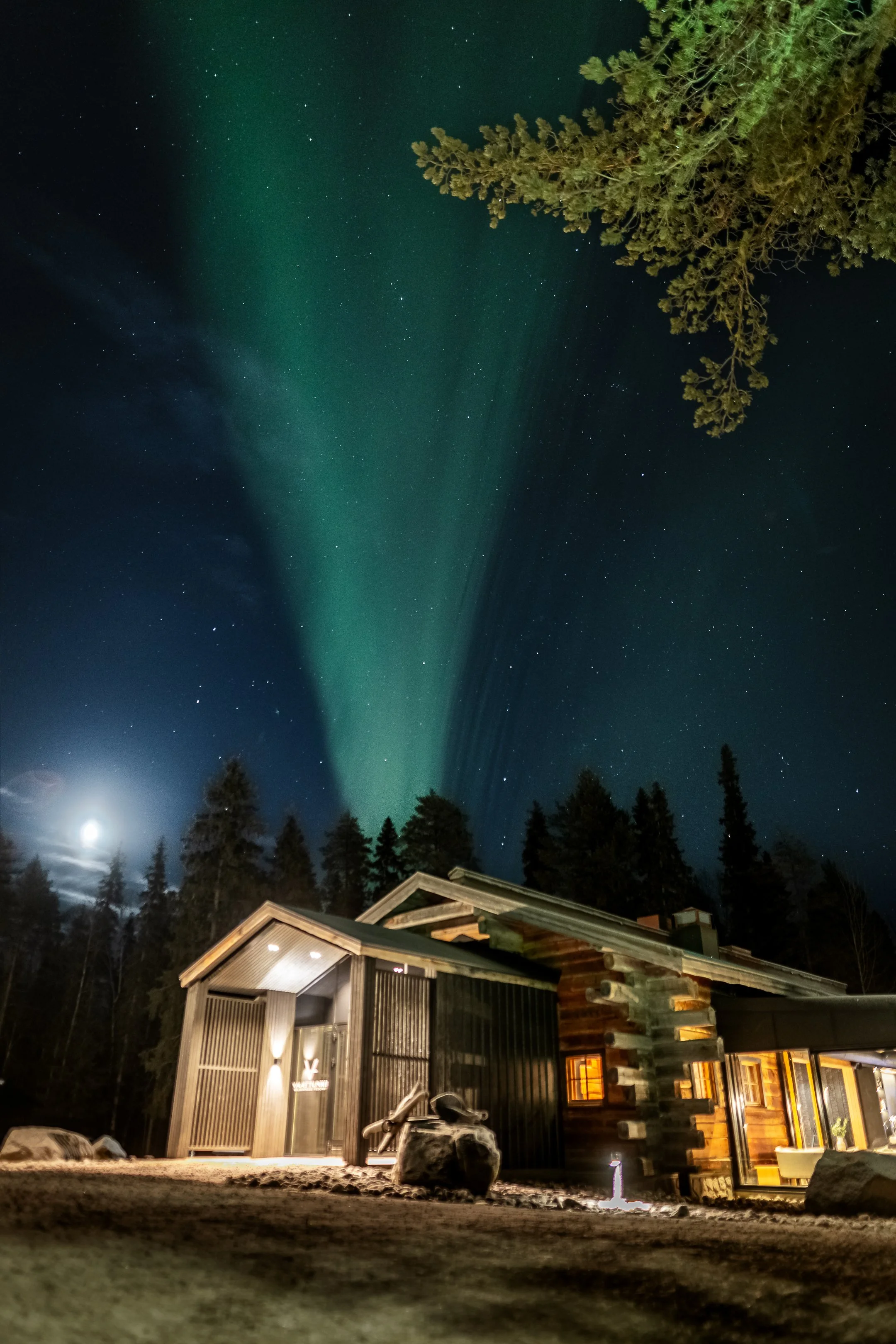 Yötöinen maisema, jossa vuoristoinen taivas ja revontulia, täysikuu ja eristävä talo, ympäröi metsä