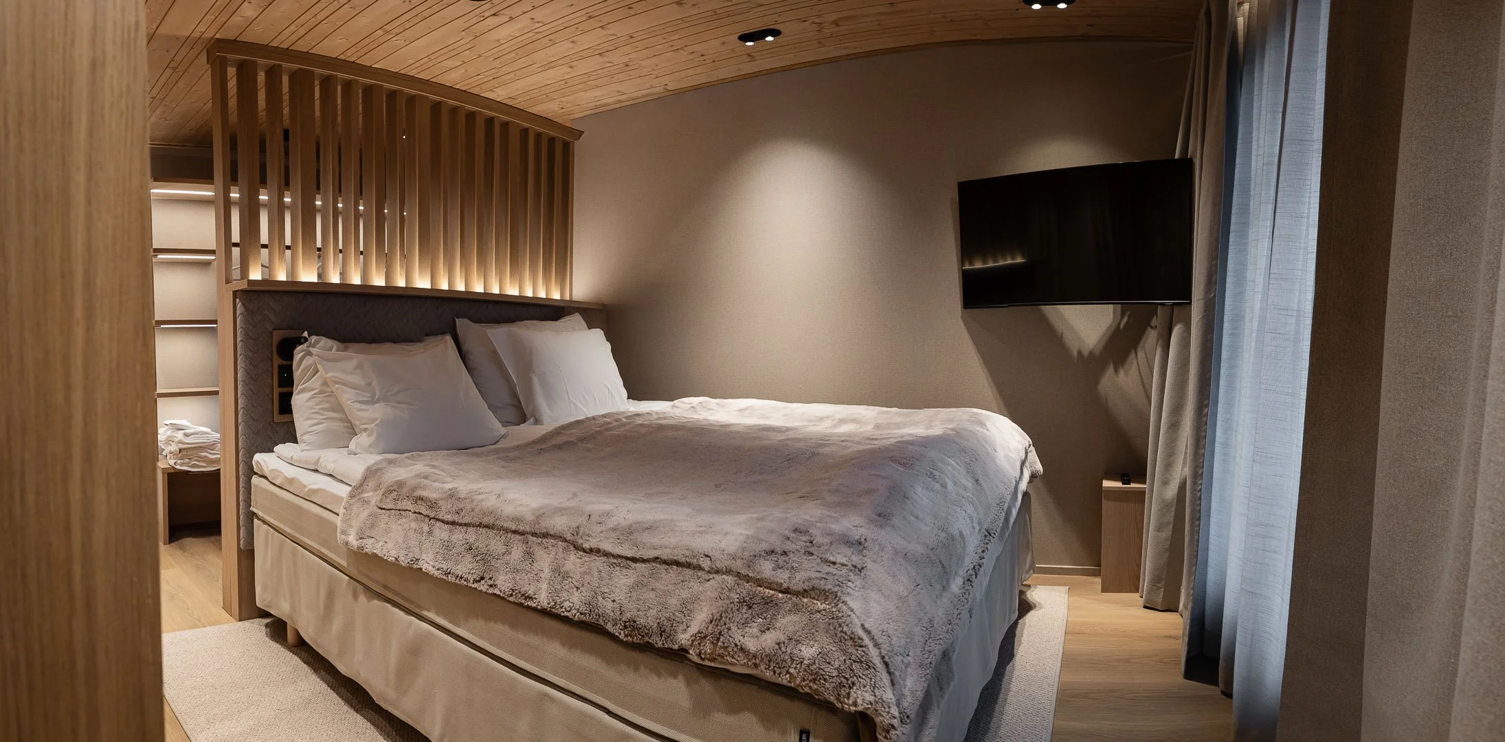 Moderni makuuhuone, jossa suuri sänky pehmeällä peitolla ja useilla valkoisilla tyynyillä, puinen seinäkiila ja raita aiheinen sisustus, seinä televisio ja tilaa luonnonvalolle suuresta ikkunasta, jossa on harmaat verhot.