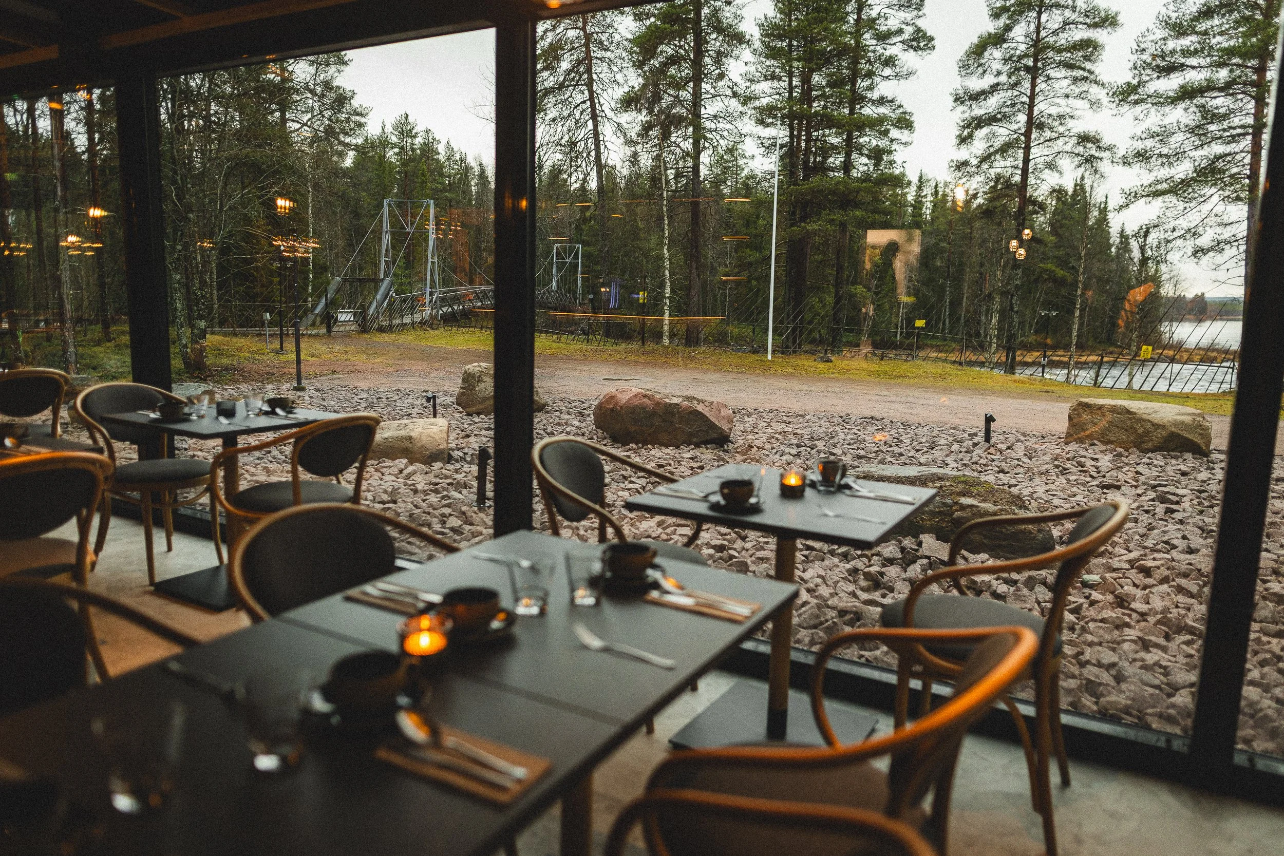 Ravintolan sisätilat ja suuret ikkunat, jotka tarjoavat näkymän ulos metsään ja vesistöön. Pöytä on asetettu ruokailua varten, kynttilä ja astioita.