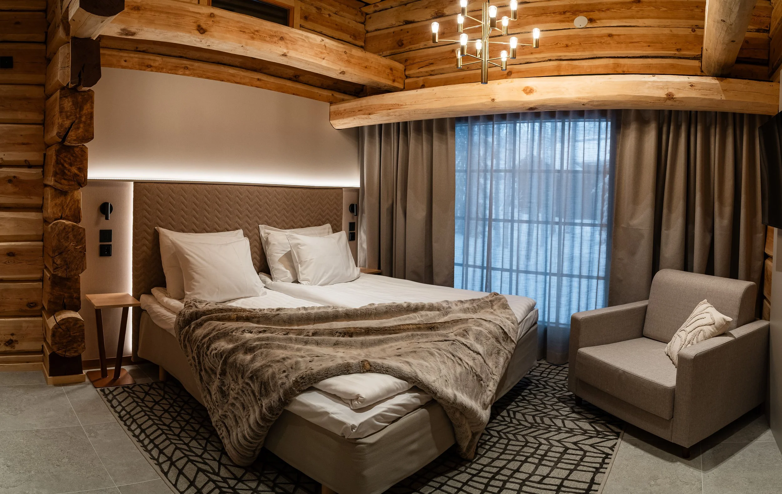 Kuvassa moderni makuuhuone, jossa on suuri sänky, pehmeitä tyynyjä, harmaita verhoja ja ilmainen näköala ikkunasta, rustiikkisia puutalojen seinäkiinnikkeitä ja kattoja.