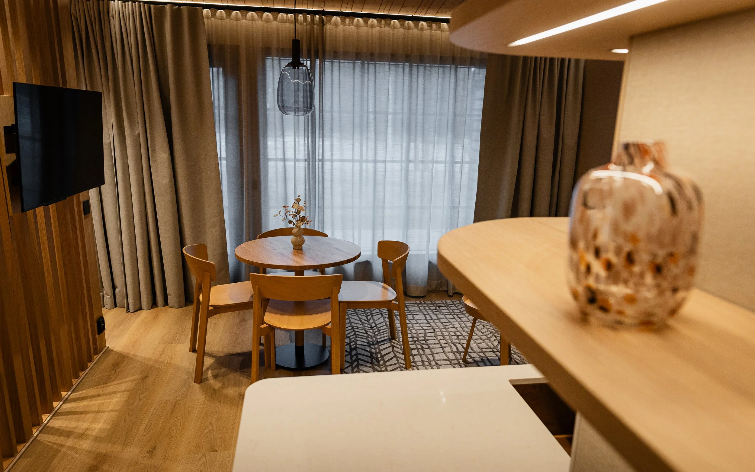 Sisustuskuva, jossa on pyöreä puinen ruokapöytä ja neljä tuolia, sekä suuri ikkuna verhoiltu harmailla ja valkoisilla verhoilla. Taustalla on seinä ja katto, jossa on piilovalot ja lamppu, sekä seinälle ripustettu televisio.