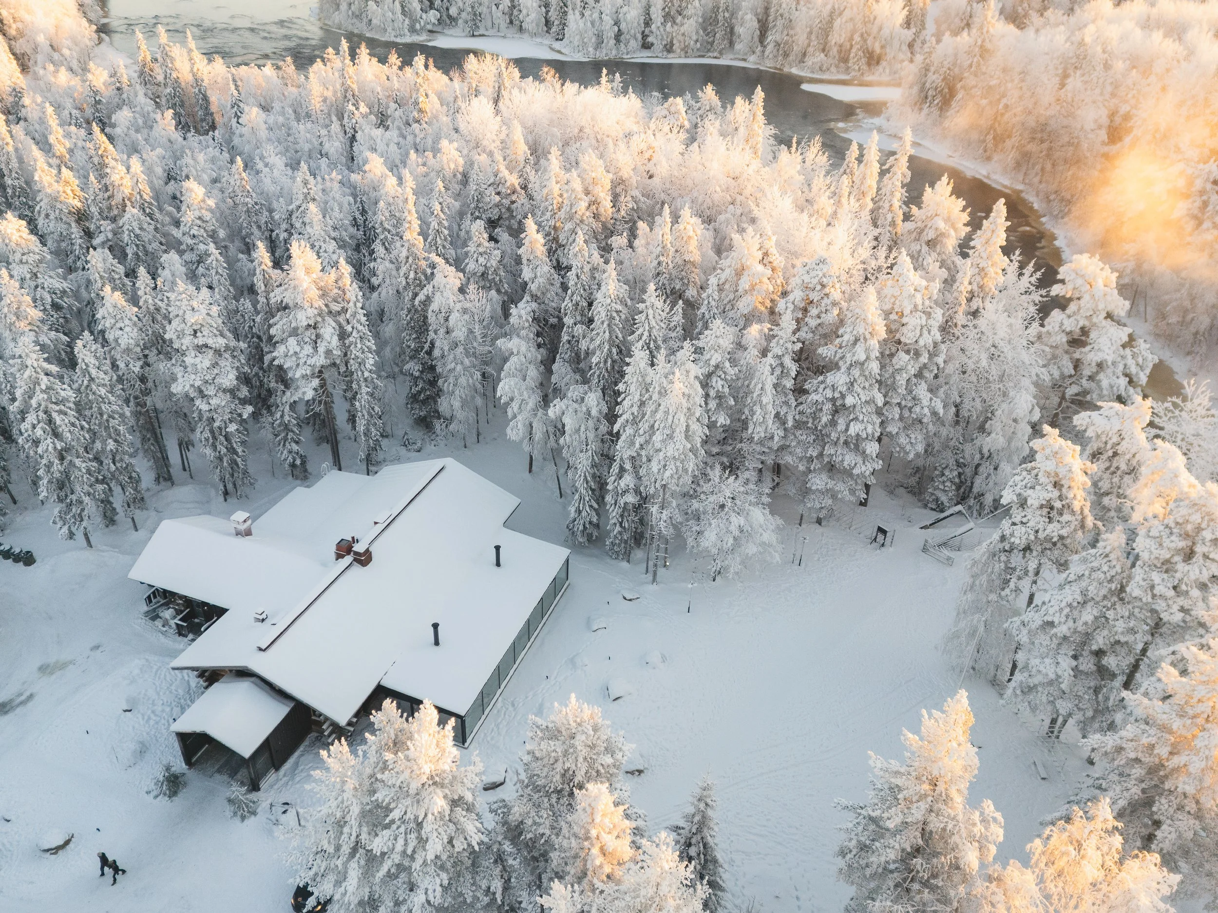 Lentosilmäkuva lumisesta mökistä ja metsistä, joki taustalla.