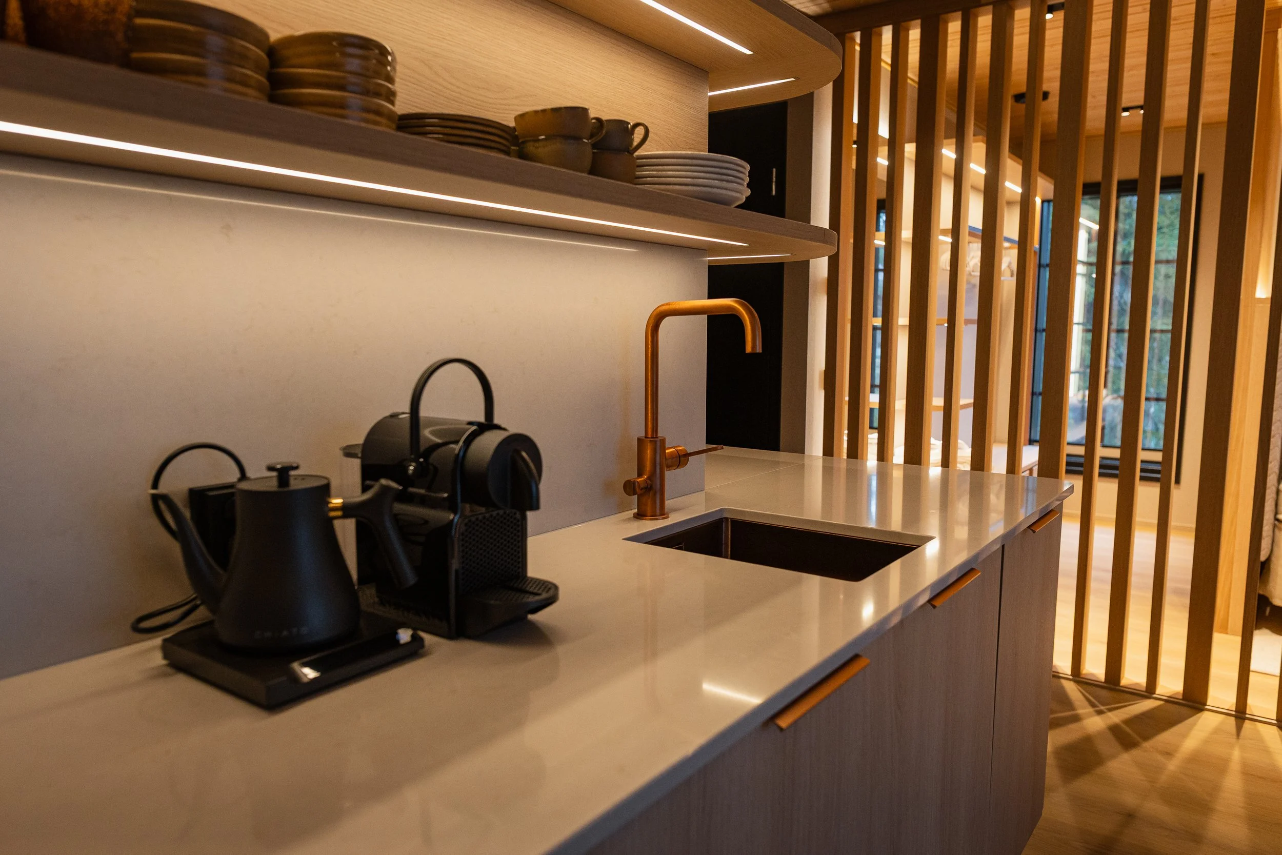 Keittiöalue, jossa on kahvikone, vieressä kupari-viemäröity hana ja jätteellinen allas. Taustalla hylly ja astioita, puiset parvekkeet.