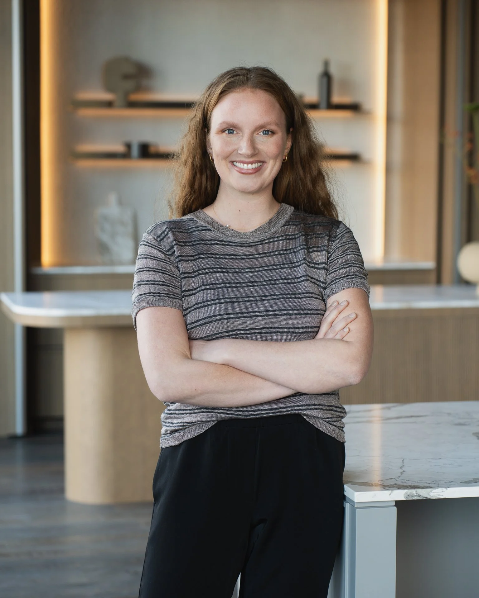 A woman with long, curly red hair, smiling, standing with her arms crossed in a modern, well-lit interior space with shelves and decorative items in the background.