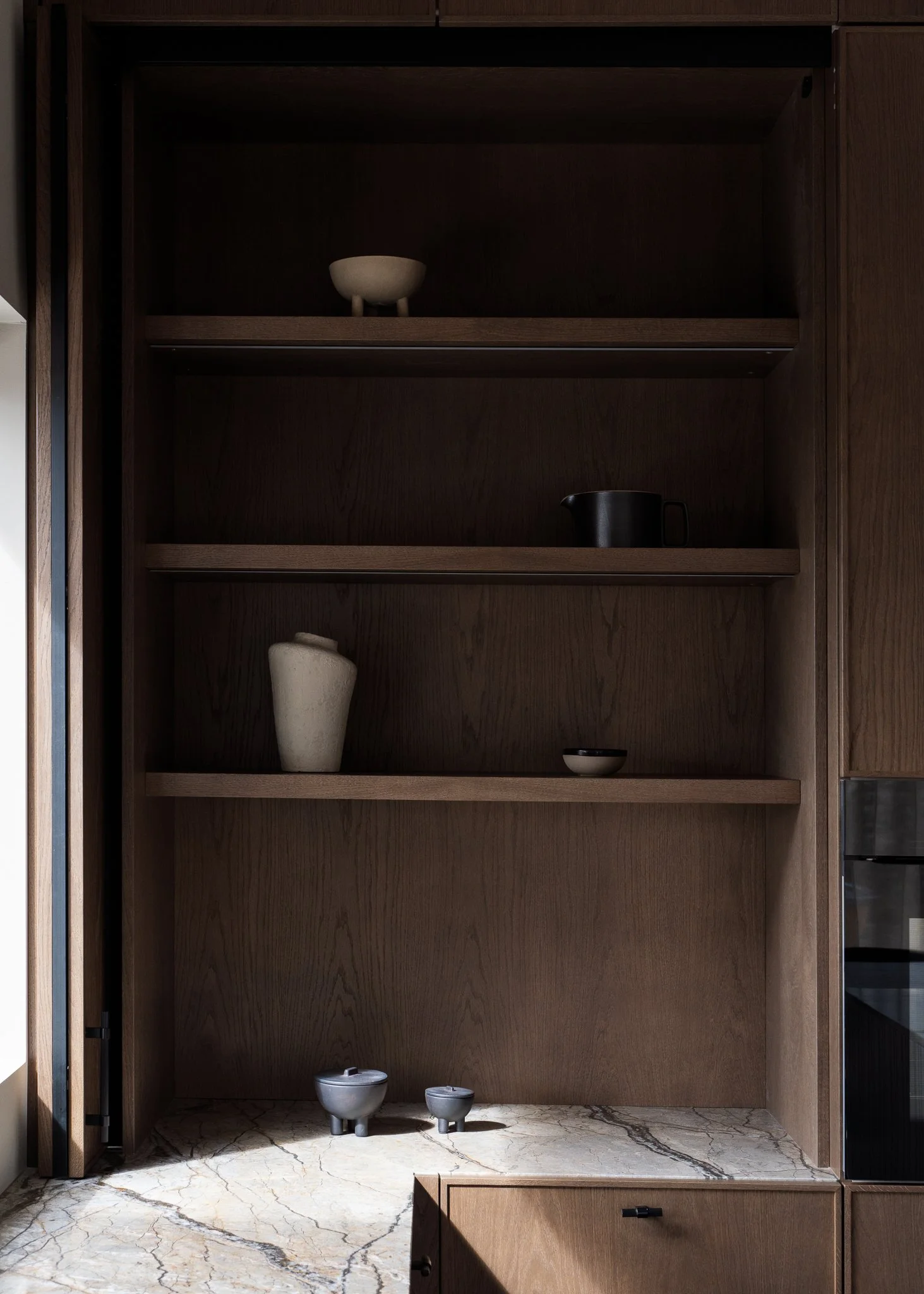 Empty wooden cabinet with three shelves, containing a white bowl with three legs on the top shelf, a black pitcher on the second shelf, and a stacked set of white bowls and two small white containers with lids on the marble countertop.