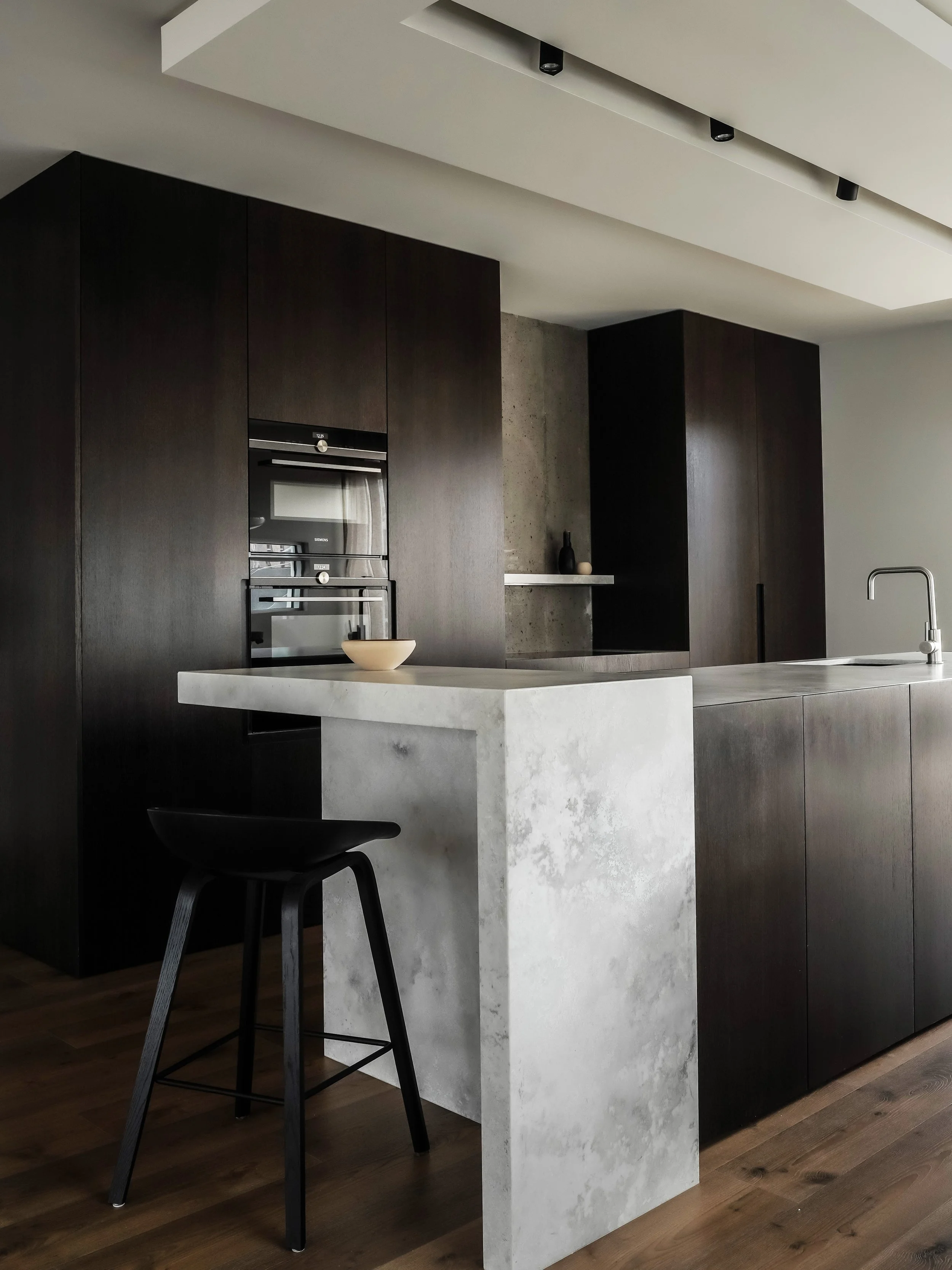 Modern kitchen with dark wood cabinets, a concrete island with a beige bowl, and black bar stool.