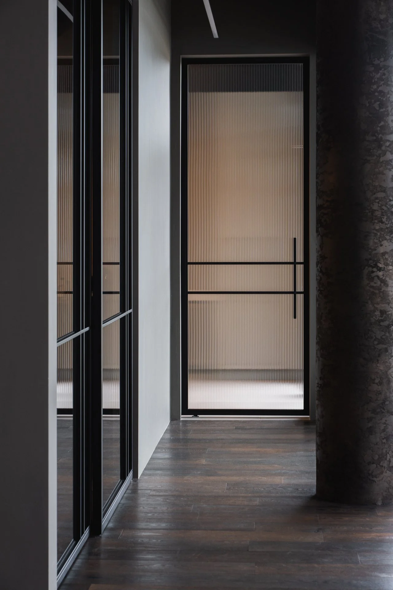 Modern industrial hallway with dark wood flooring, black metal-framed glass closet doors on the left, and a frosted glass door at the end of the corridor.