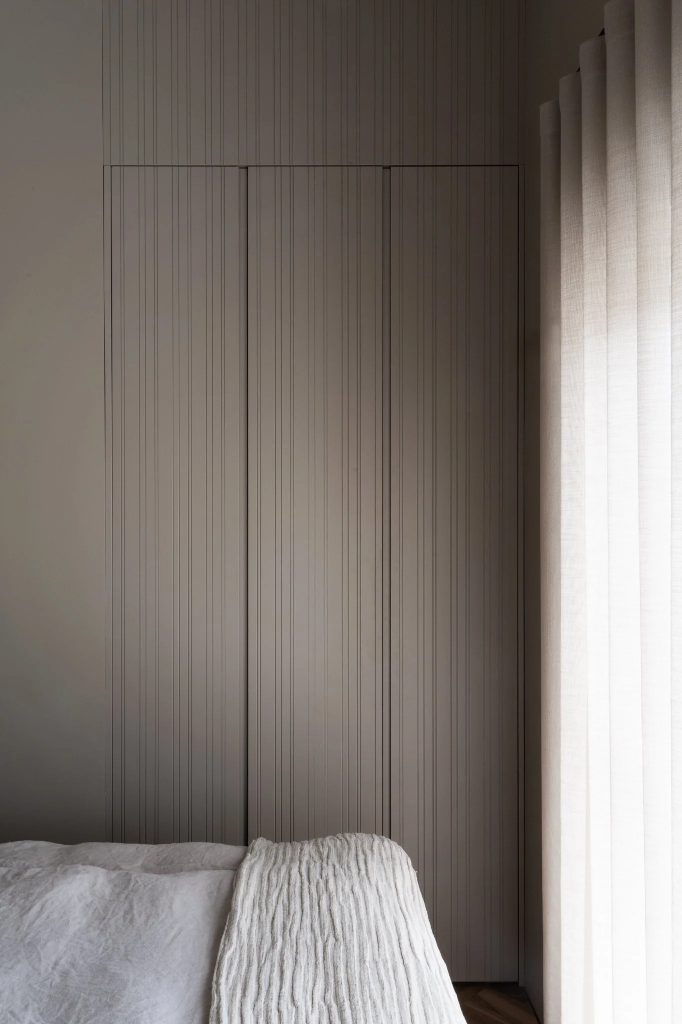 View of a bedroom with a bed covered in a light, textured blanket and a pillow, a wooden paneled wall, and sheer curtains on the window.