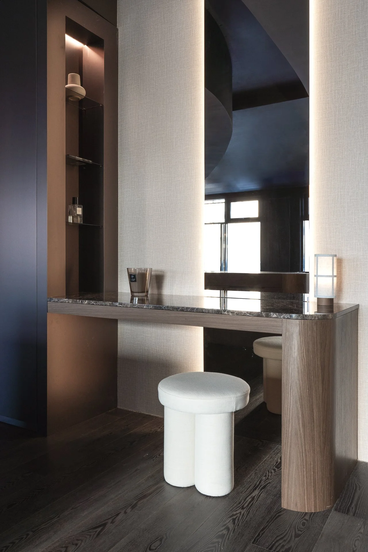 Modern vanity area with a black marble countertop, a round white stool, and a wall with a textured beige finish. There is a large mirror, a small lamp, and a bottle of perfume on the counter.