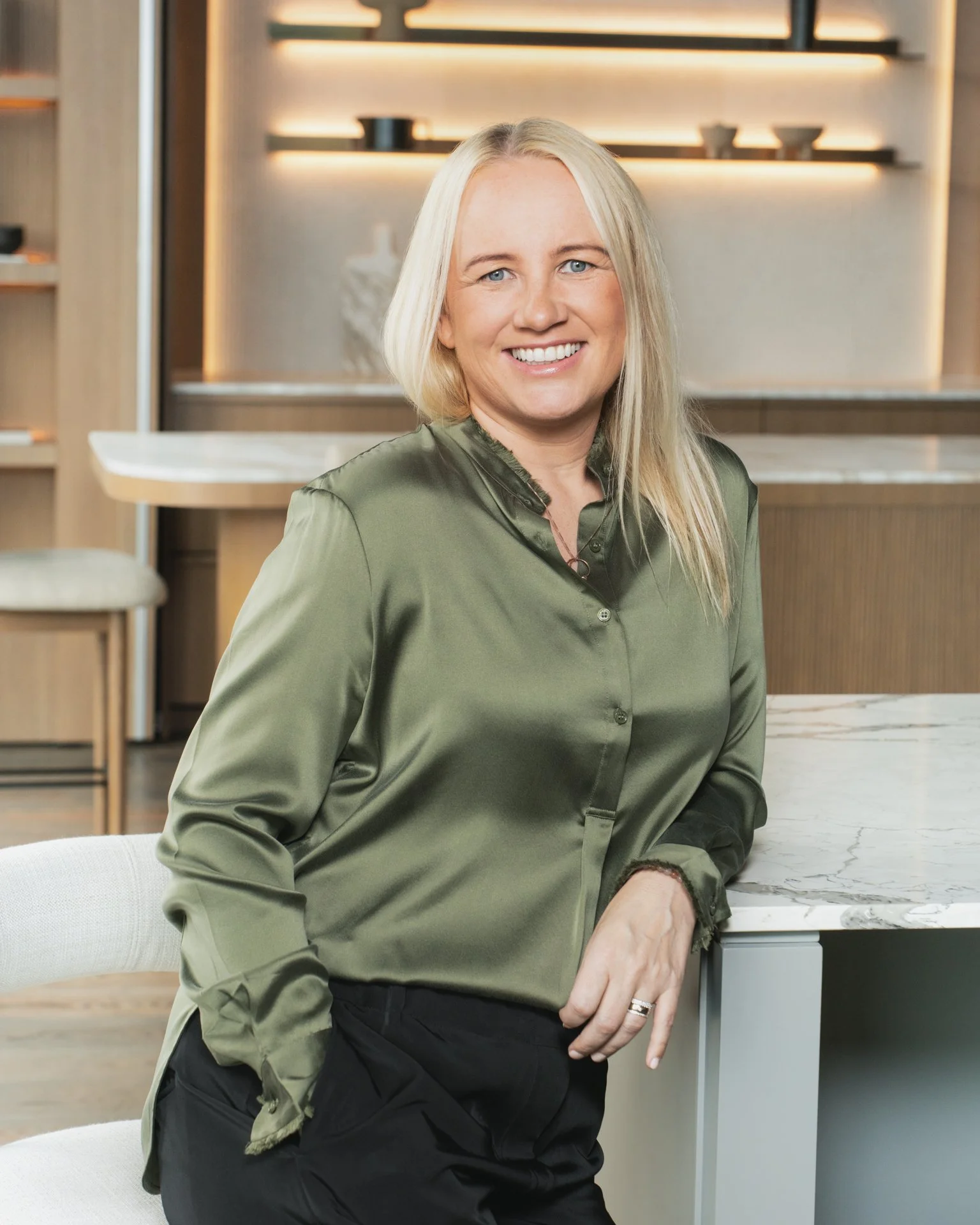 A smiling blonde woman with blue eyes wearing an olive green blouse standing in a modern kitchen with wooden shelves and white marble countertop.