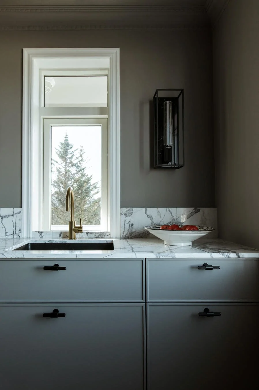 Modern kitchen sink with a gold faucet, white marble countertop, and a bowl of tomatoes, with a window showing trees outside.