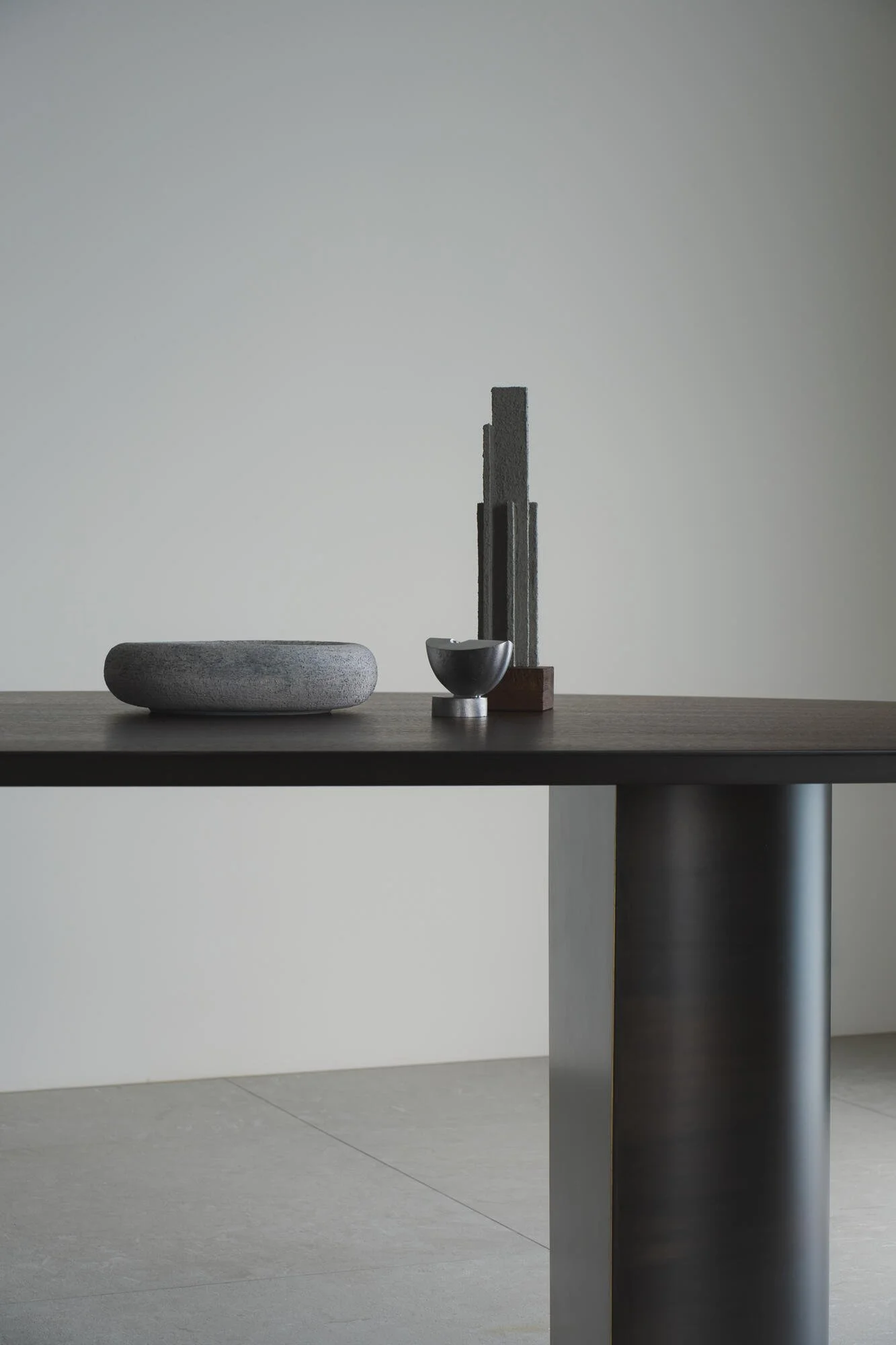 Minimalist interior with a sleek black table, a large textured stone bowl, a small metallic bowl, and modern architectural sculptures against a plain white wall.