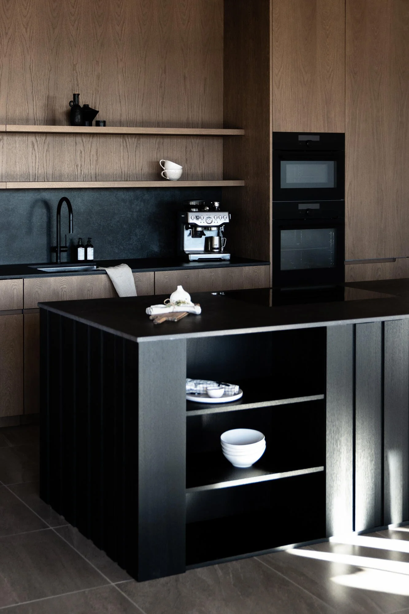 Modern kitchen with wooden cabinets, black countertops, a coffee machine, open shelving with cups and bottles, and a black and gray kitchen island with open shelves containing white bowls and other dishes.