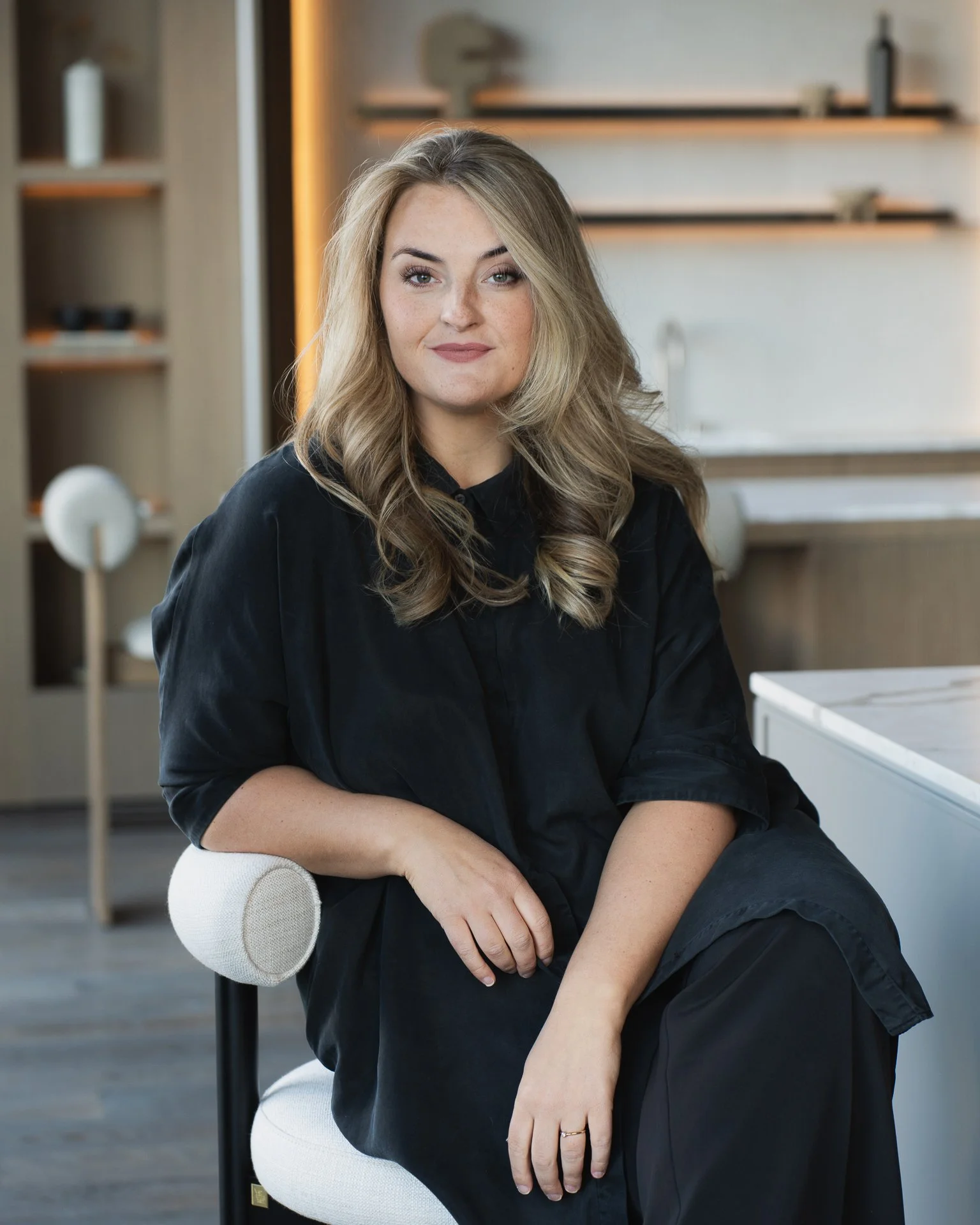 A woman with long blonde hair, wearing a black shirt, sits casually in a modern, well-lit room. She has a relaxed posture, with one arm resting on her knee and a slight smile on her face.