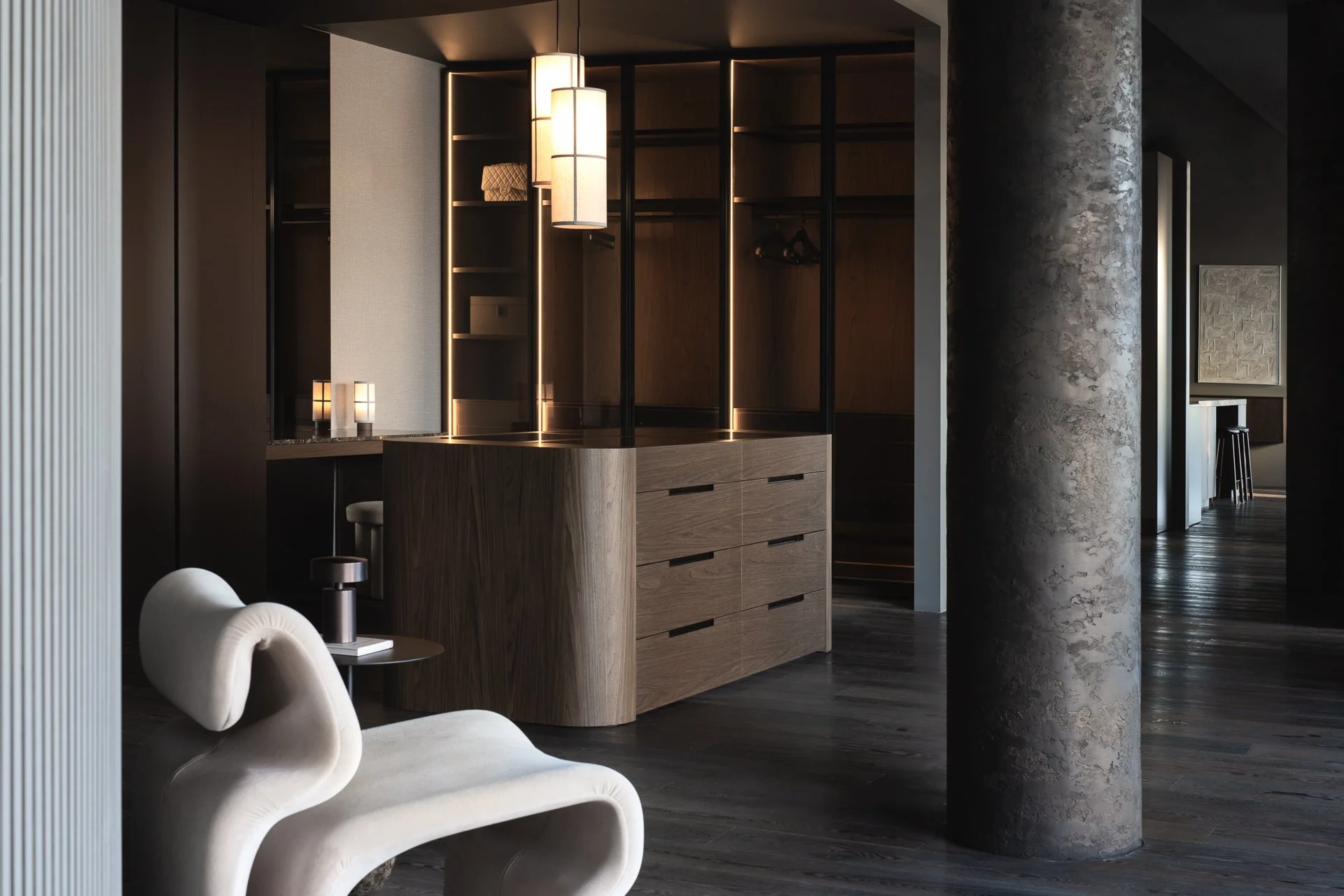 Modern interior space with a wooden cabinet, a textured gray concrete column, and abstract white chairs in the foreground. Hanging pendant lights illuminate the area.