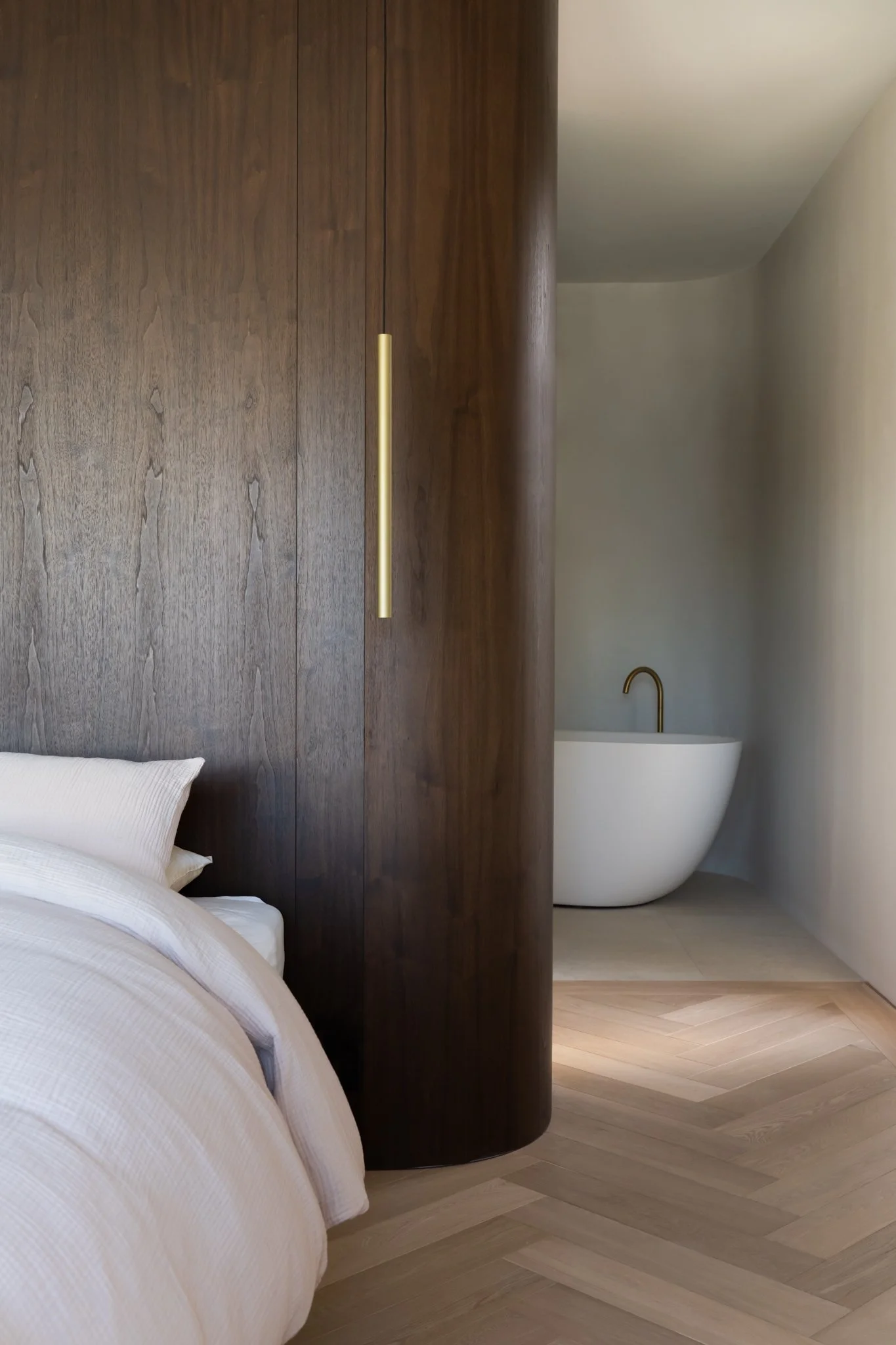 A bedroom with a wooden partition, a bed with white bedding, and a bathroom with a white freestanding bathtub and a black faucet in the background.