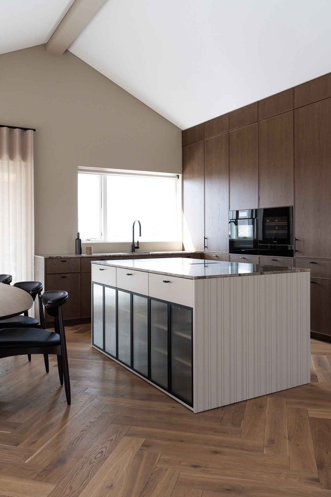 Modern kitchen with wood cabinets, a kitchen island with a white base and dark glass-paneled sides, a large window, and a wooden floor.