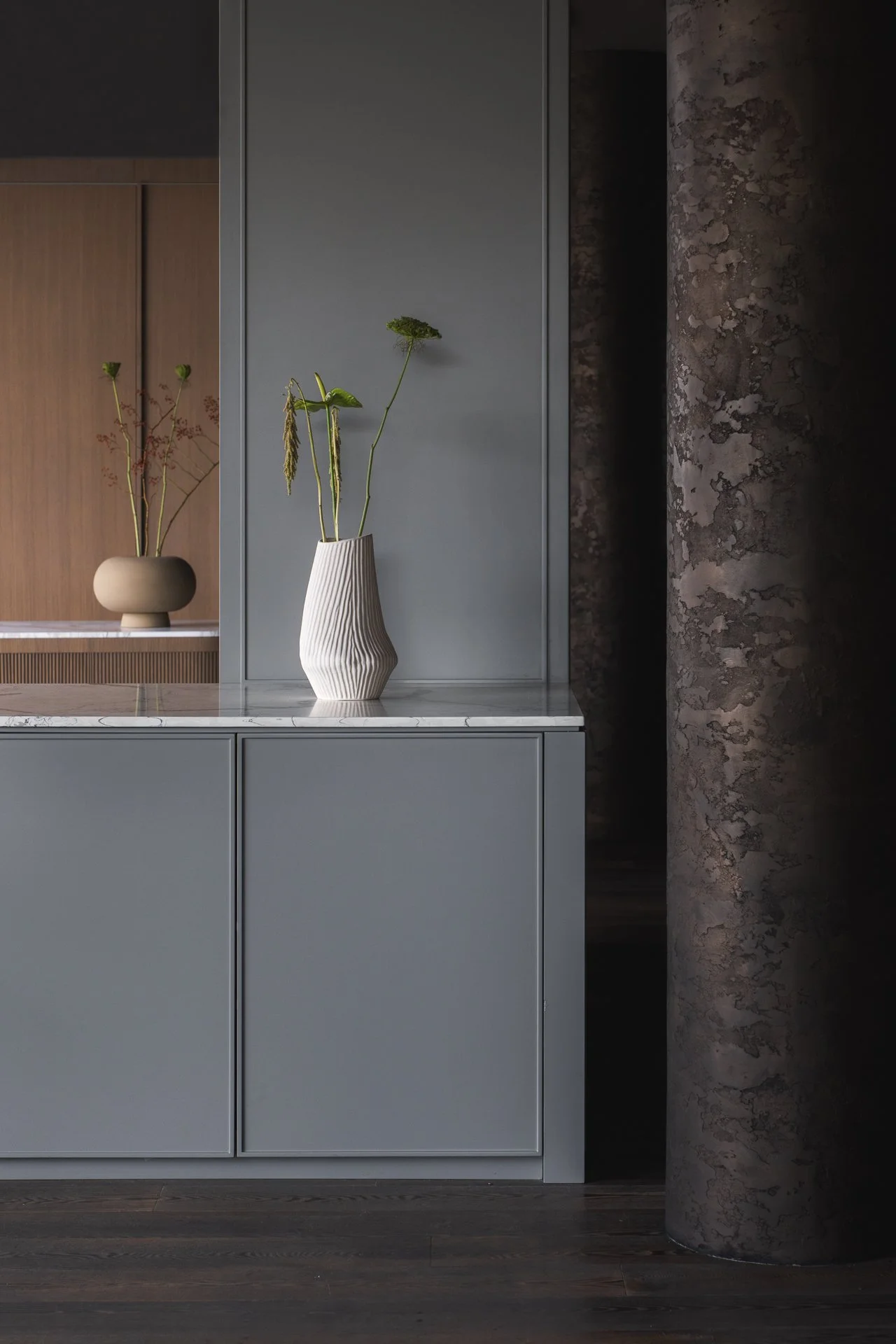 Minimalist interior with a light blue cabinetry, a gray wall, and a marble countertop. Two vases with green plants are on the counter. A wooden panel and a decorative column are in the background.